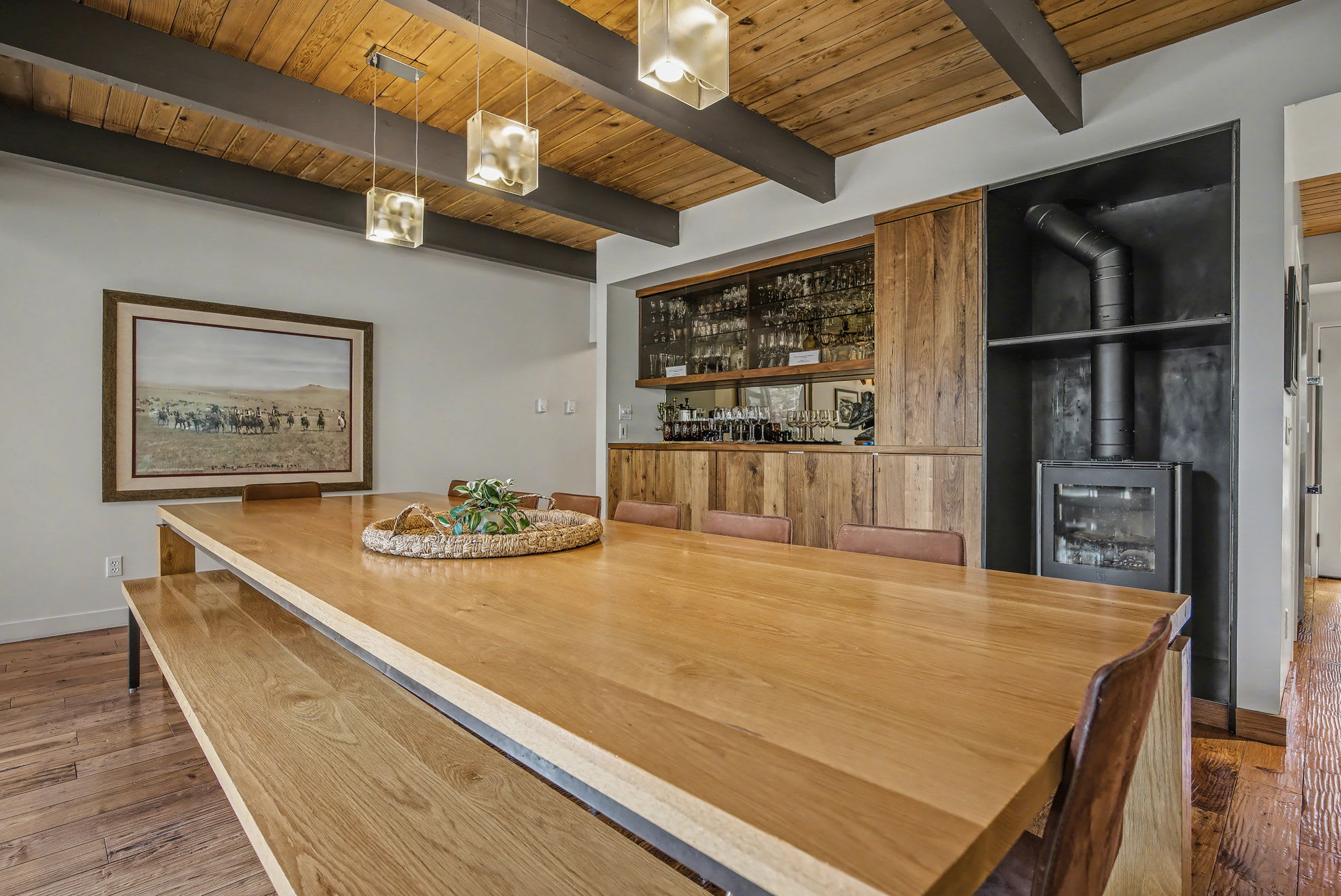 A grand living room features a central stone fireplace and floor-to-ceiling windows offering panoramic views of the surrounding forest. Multiple dark sofas and armchairs are arranged around a large coffee table under a vaulted wooden ceiling.