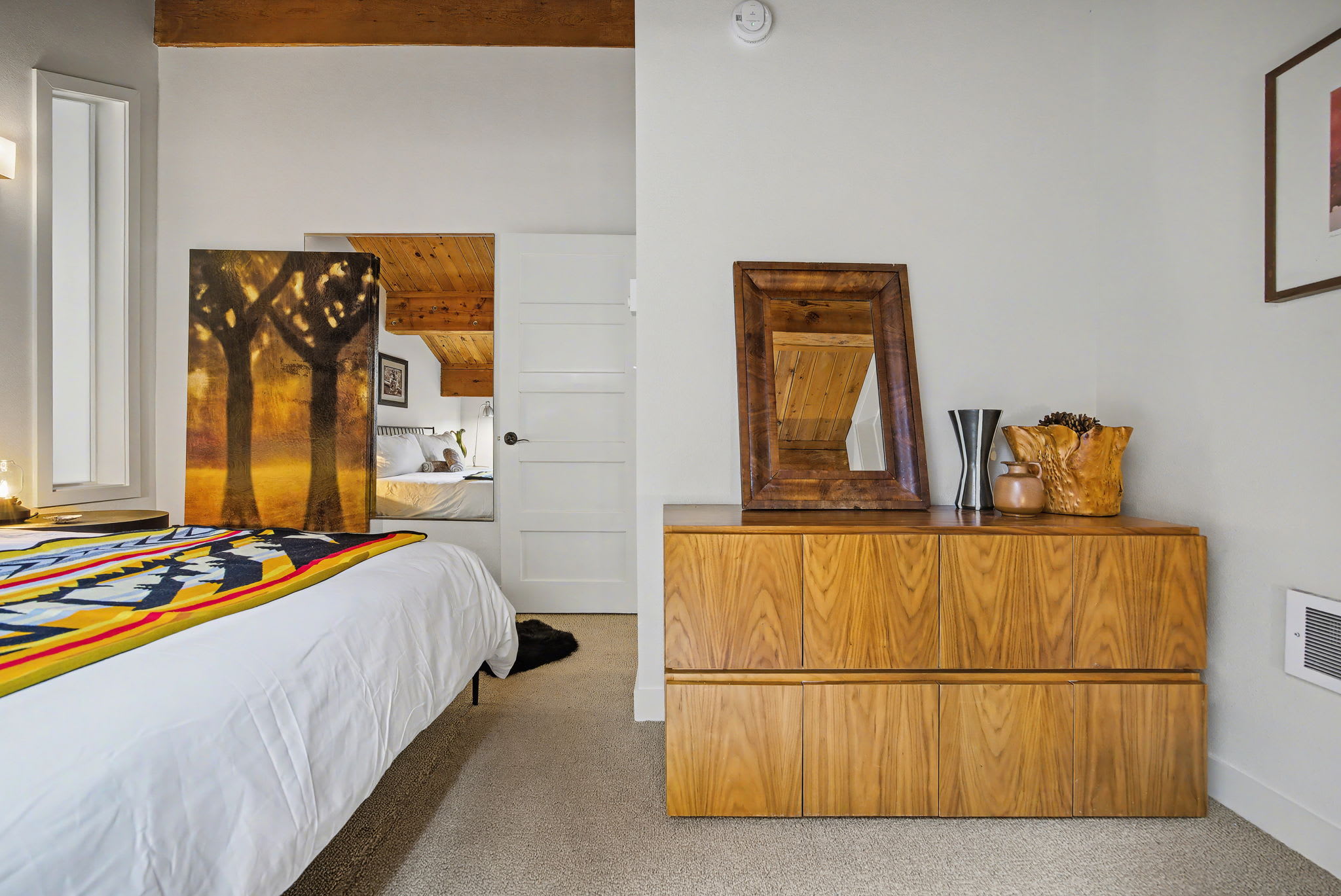 A cozy bedroom features a queen-sized bed with crisp white bedding and a vibrant, colorful patterned blanket, flanked by dark round nightstands. Exposed wooden beams accent the vaulted ceiling, and framed artwork adorns the white walls above a soft carpet.