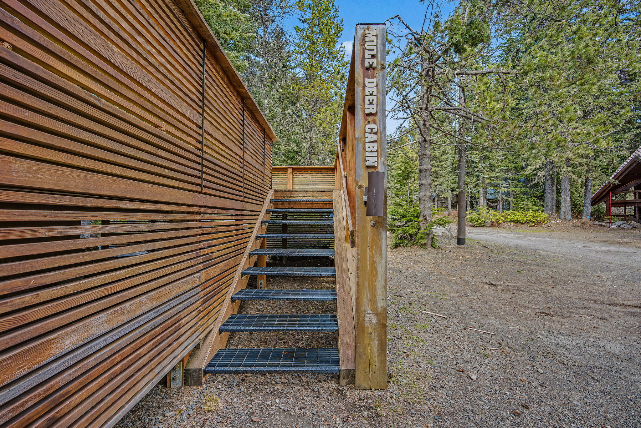 The front exterior of the chalet, showcasing its stone and wood construction amidst a dense forest. A slatted wooden fence with a large white deer head cutout provides privacy and a distinctive entrance.