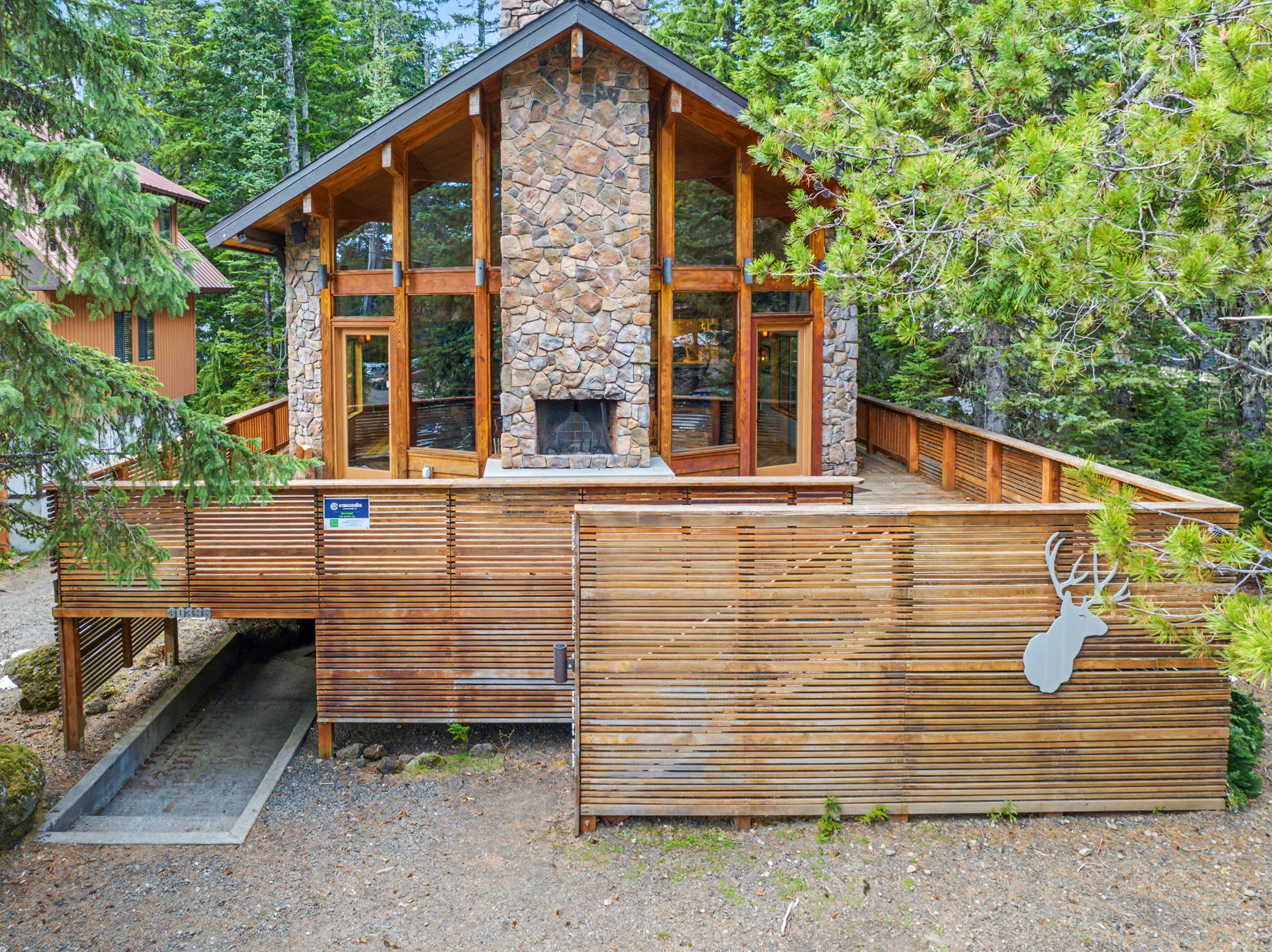 The front exterior of a rustic house, featuring a prominent stone facade with large windows and a central stone chimney. A spacious wooden deck wraps around the front, surrounded by lush green trees.