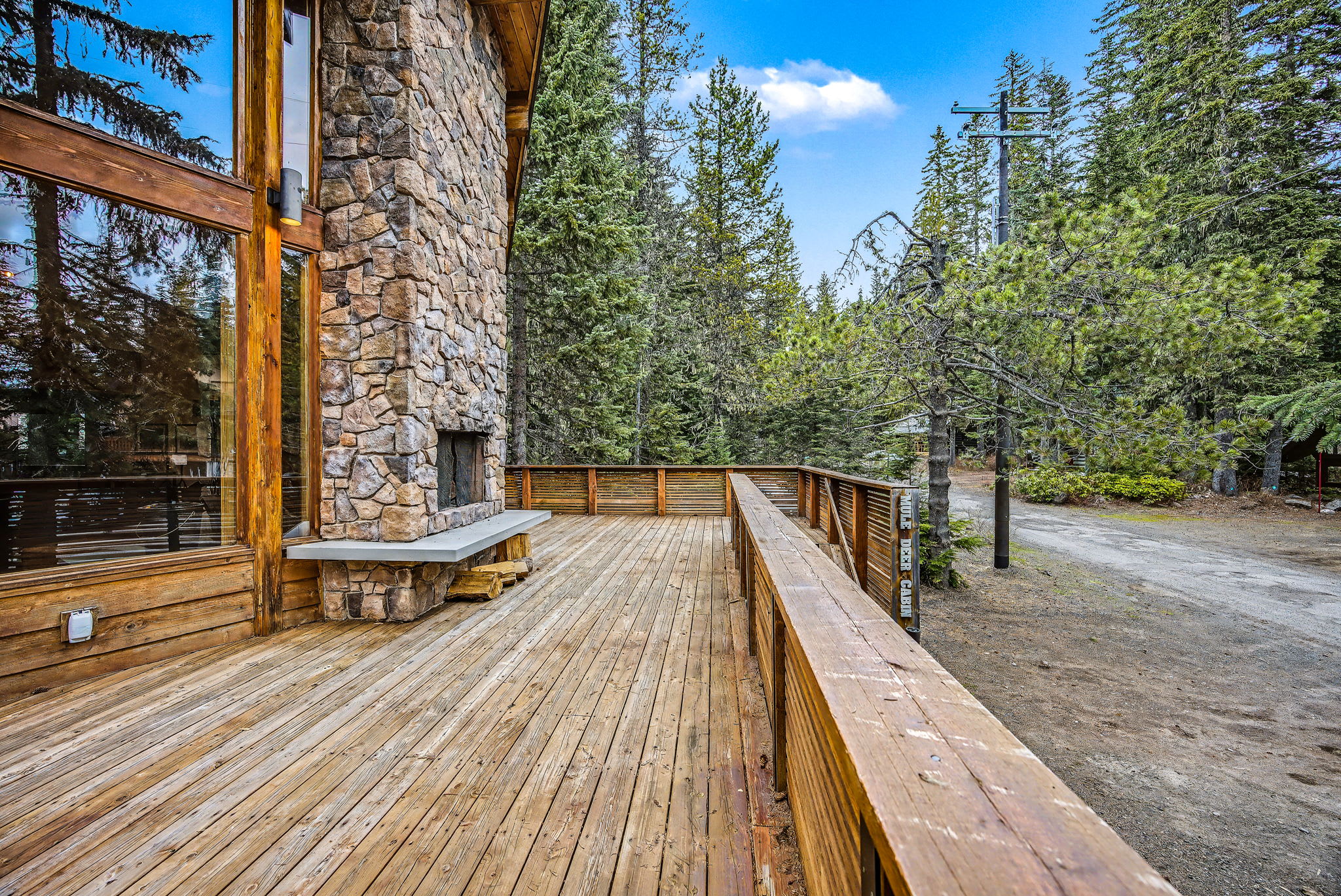 A wide wooden deck extends from the house, featuring a built-in stone fireplace and offering views of the surrounding pine forest and a dirt road. The deck has a slatted wooden railing.