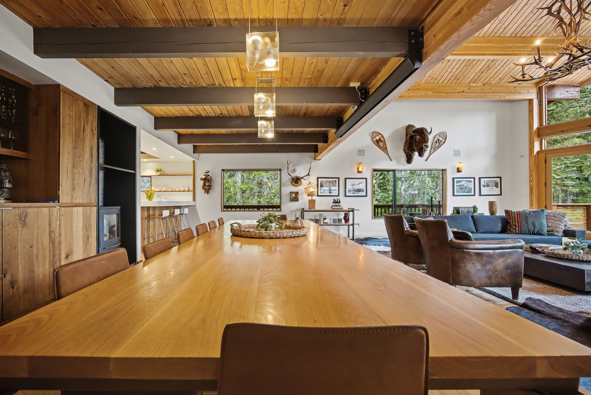This wide view of the living room highlights a large dark sectional sofa, a wooden coffee table, and a wall decorated with a buffalo head mount and snowshoes. Large windows provide a lush green outdoor vista, enhancing the rustic-modern style.