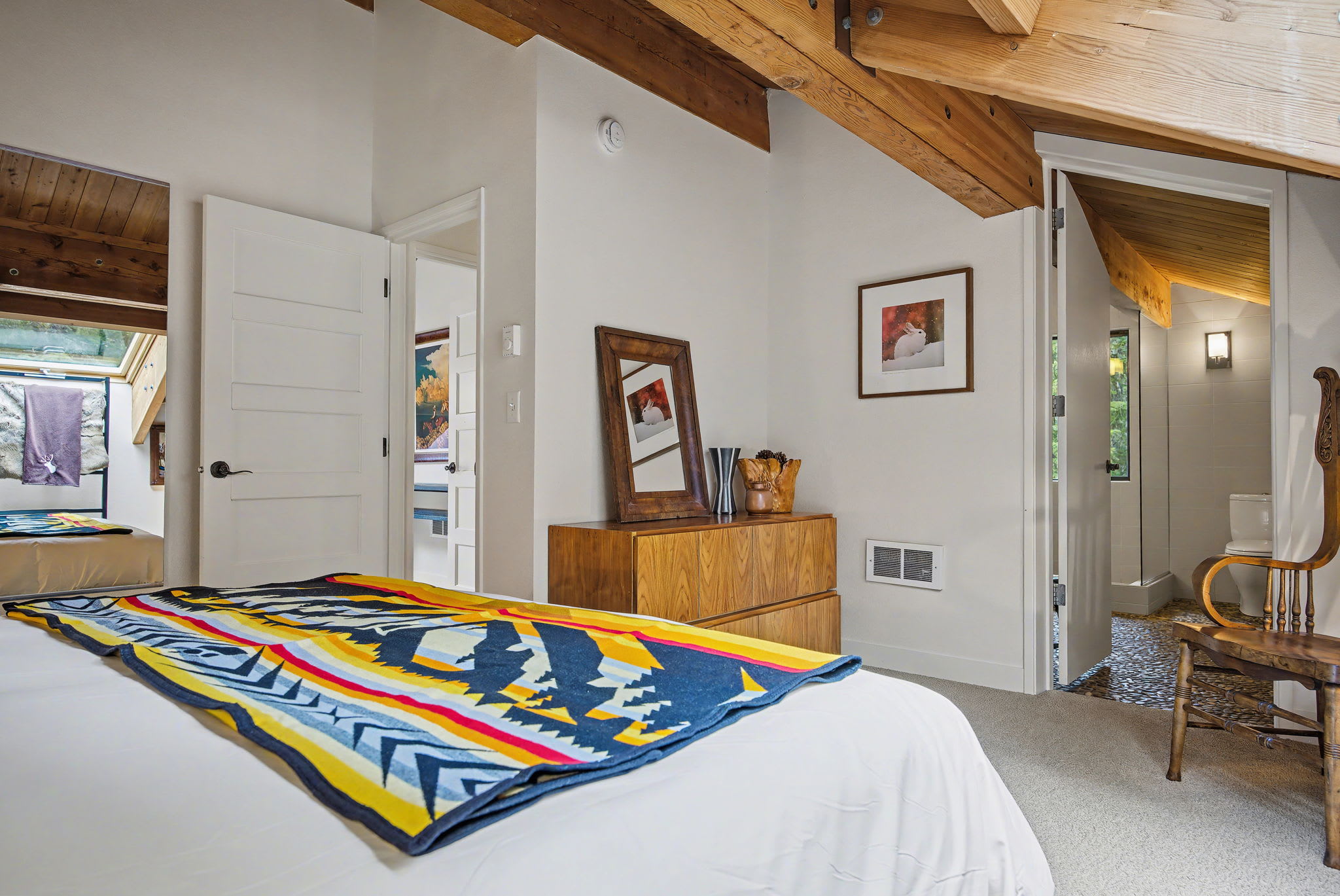 A wide shot of the bedroom showcases a queen bed with white linens and a vibrant blanket, positioned beneath a large skylight offering views of the trees. Exposed wooden beams create a rustic-modern aesthetic, complemented by framed art and a blue faux fur rug on the carpeted floor.