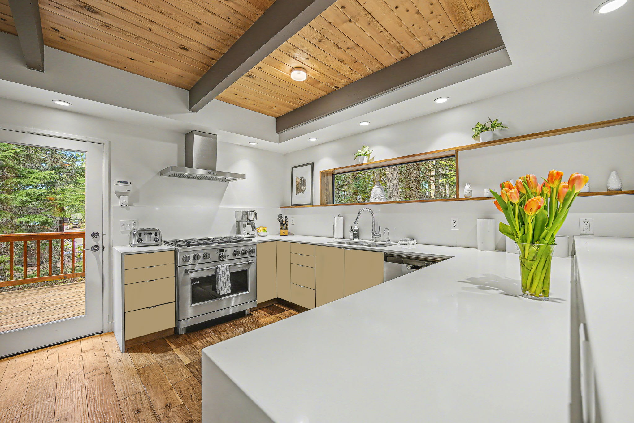 A modern kitchen with white quartz countertops, light beige flat-panel cabinets, and stainless steel appliances. A large window above the sink offers a view of the forest, and a glass door leads to an outdoor deck.