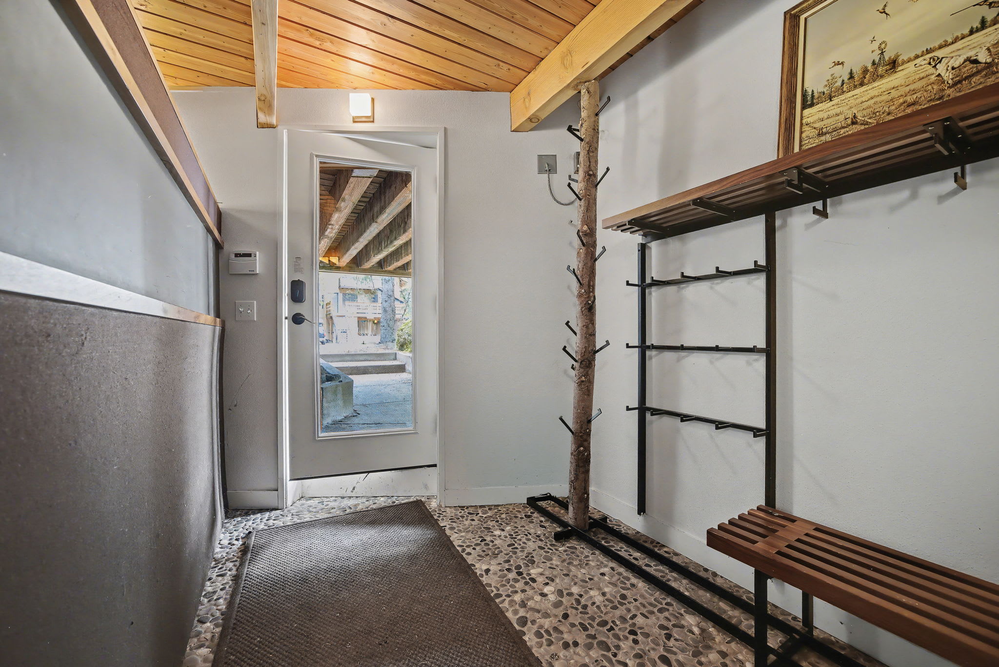 Bright entryway with a glass-paned door leading outside, featuring unique pebble flooring, a rustic tree-branch coat rack, and a wooden slatted bench for seating.