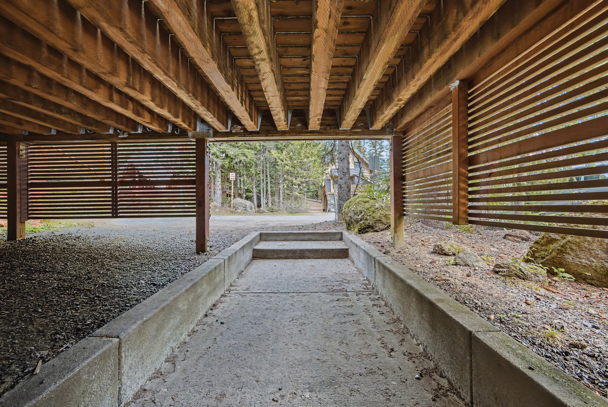 Concrete pathway leading under a large wooden deck structure with exposed beams and slatted wooden walls, surrounded by natural ground cover and distant trees.