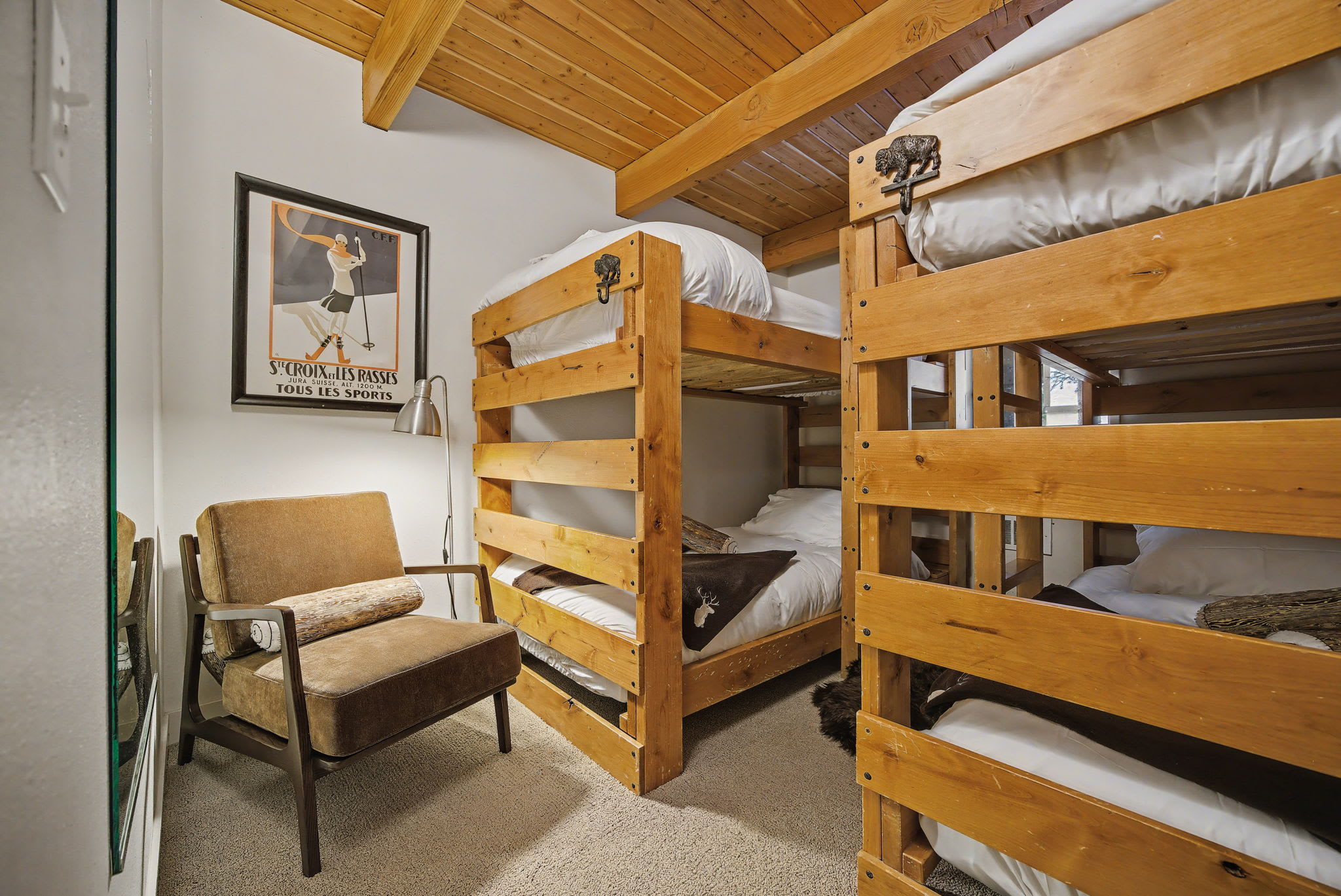 Cozy bedroom featuring wooden bunk beds with white bedding, a comfortable armchair, and a vintage ski poster on the wall, all under a wooden ceiling with exposed beams.