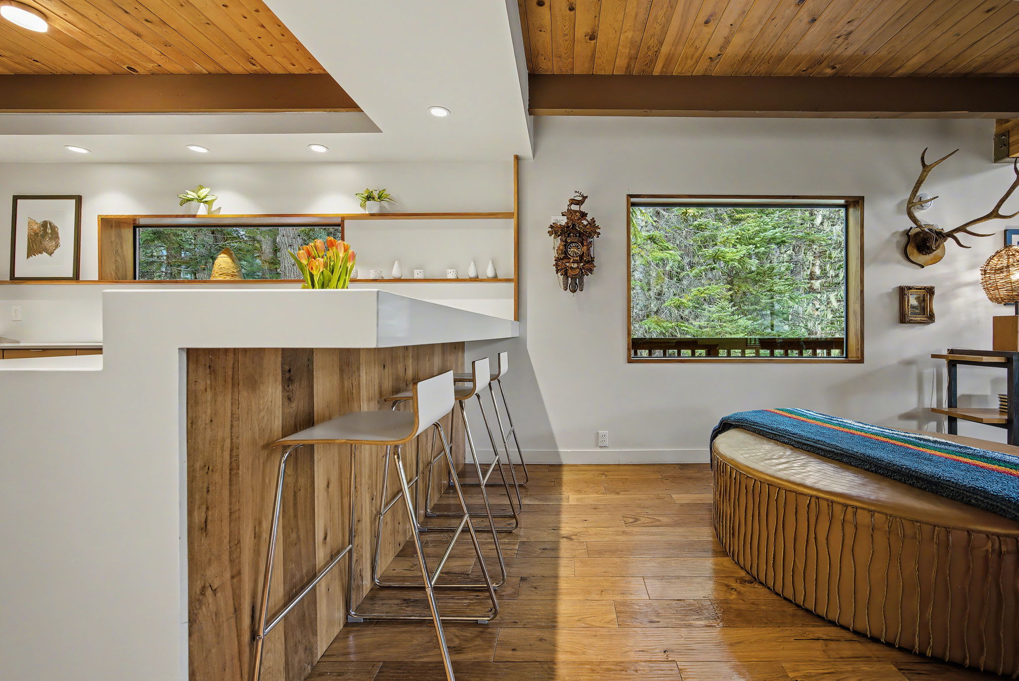 Expansive wooden deck with a gas grill, extending along the dark-stained exterior of the house, offering views of the surrounding tall evergreen trees.