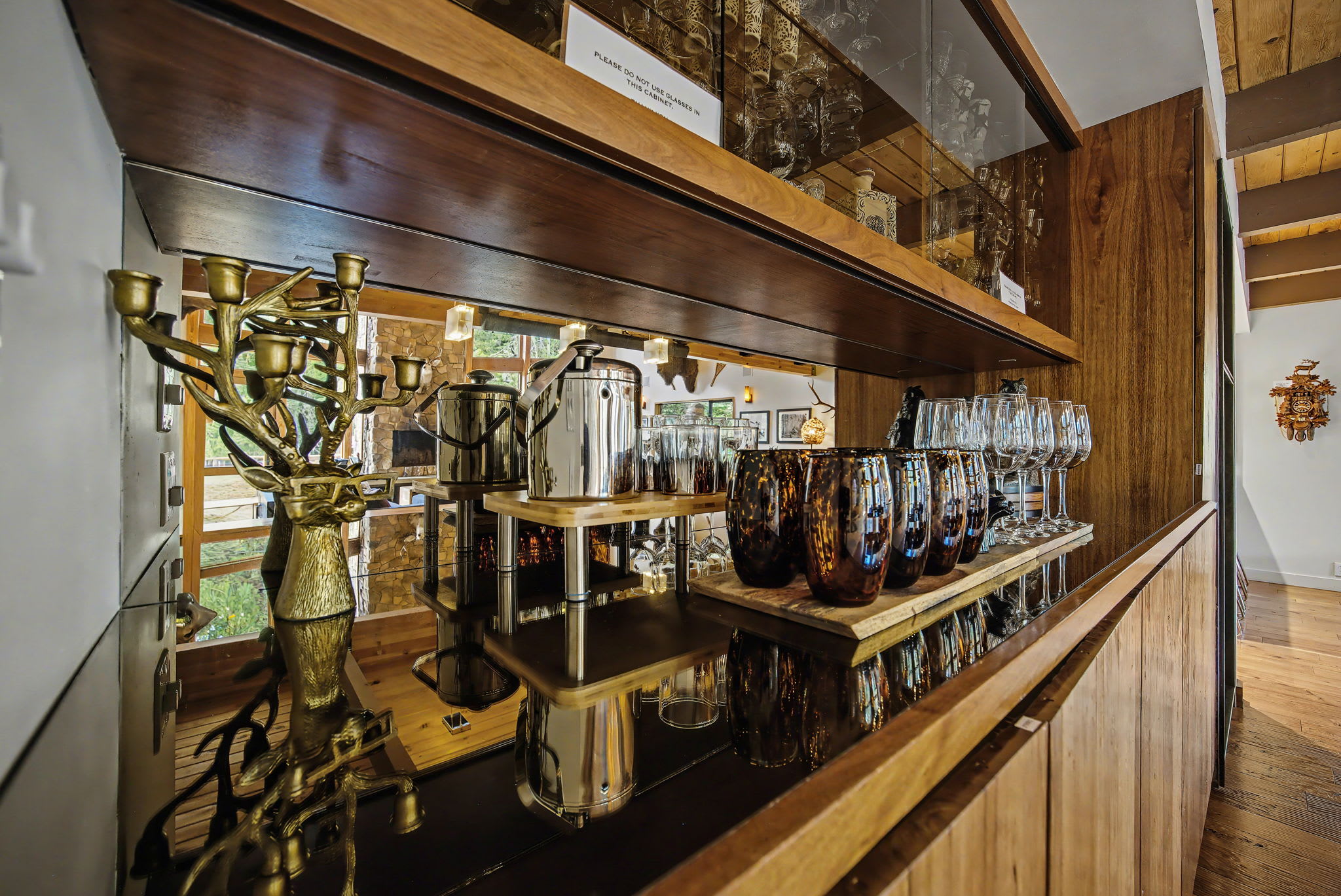 A spacious dining area features a long, light-wood dining table with benches and chairs, illuminated by modern pendant lights from a vaulted wooden ceiling. A built-in bar with mirrored shelves and a wood-burning stove are visible in the background.