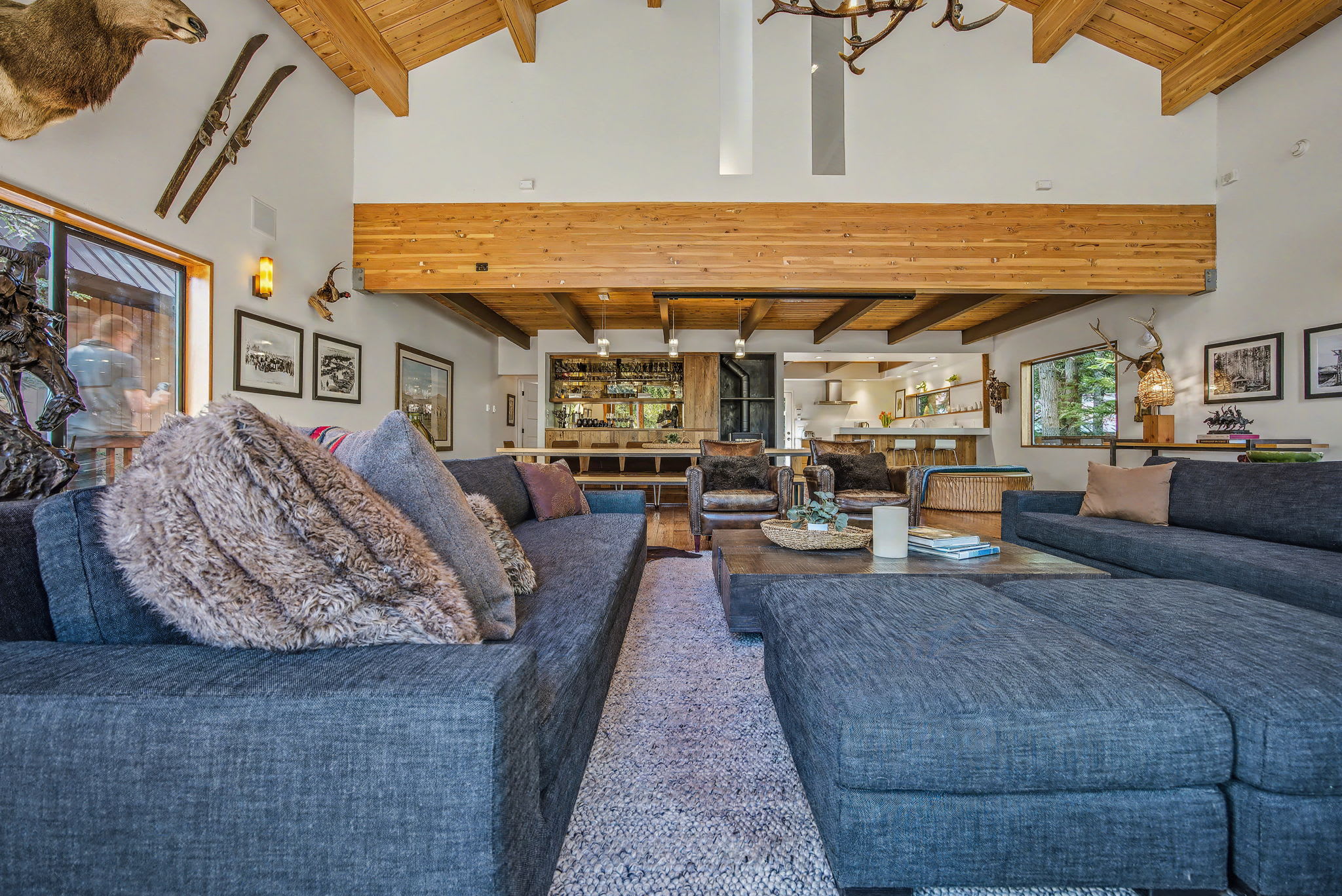 A spacious wooden deck wraps around the side of the chalet, featuring an outdoor fireplace built into the stone exterior. Large windows and a glass door connect the interior to the forest surroundings.