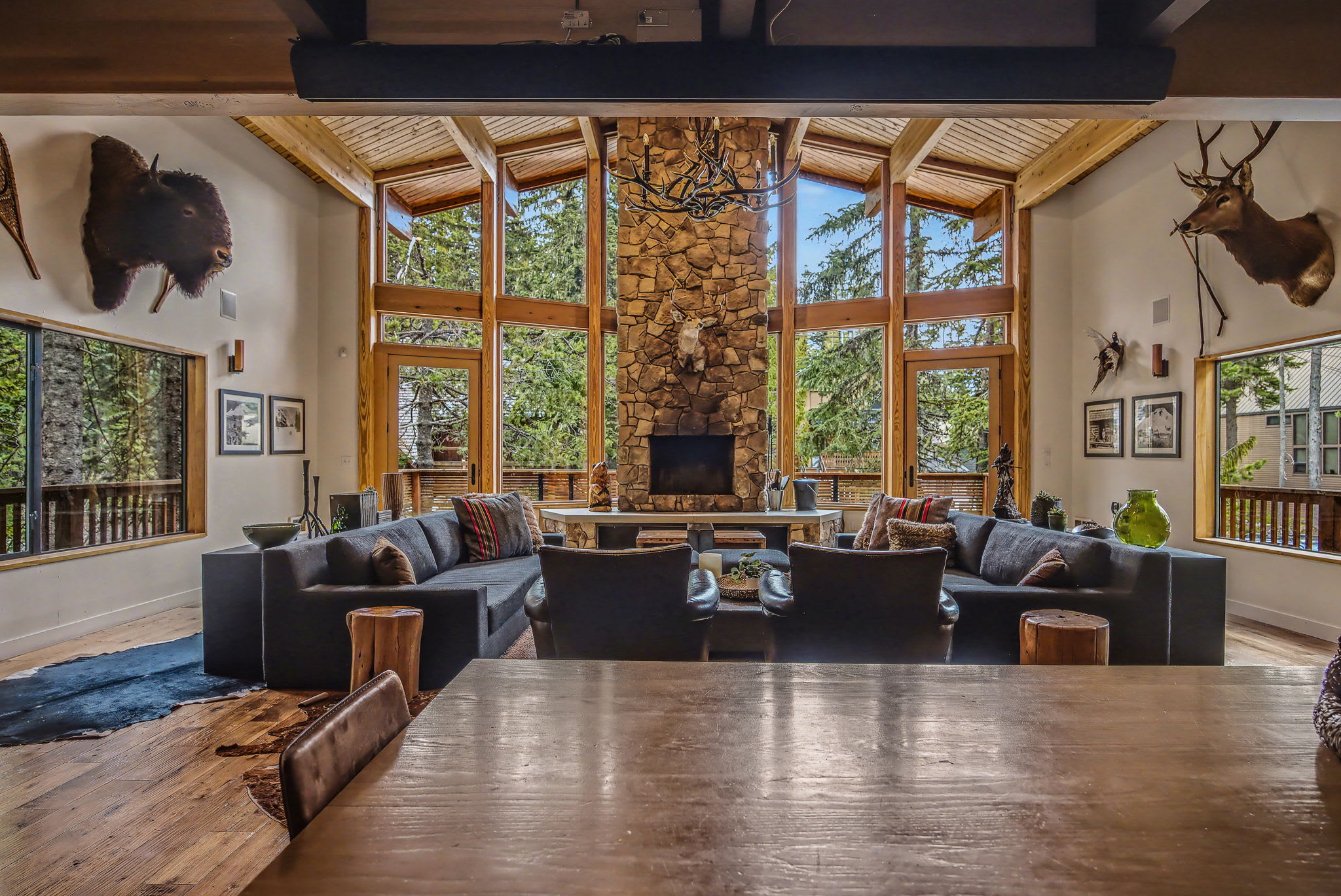 A cozy living room features a grand stone fireplace with a carved wooden bear statue, a dark sectional sofa, and a rustic wooden coffee table. Vaulted wooden ceilings and large windows offer views of the surrounding nature.