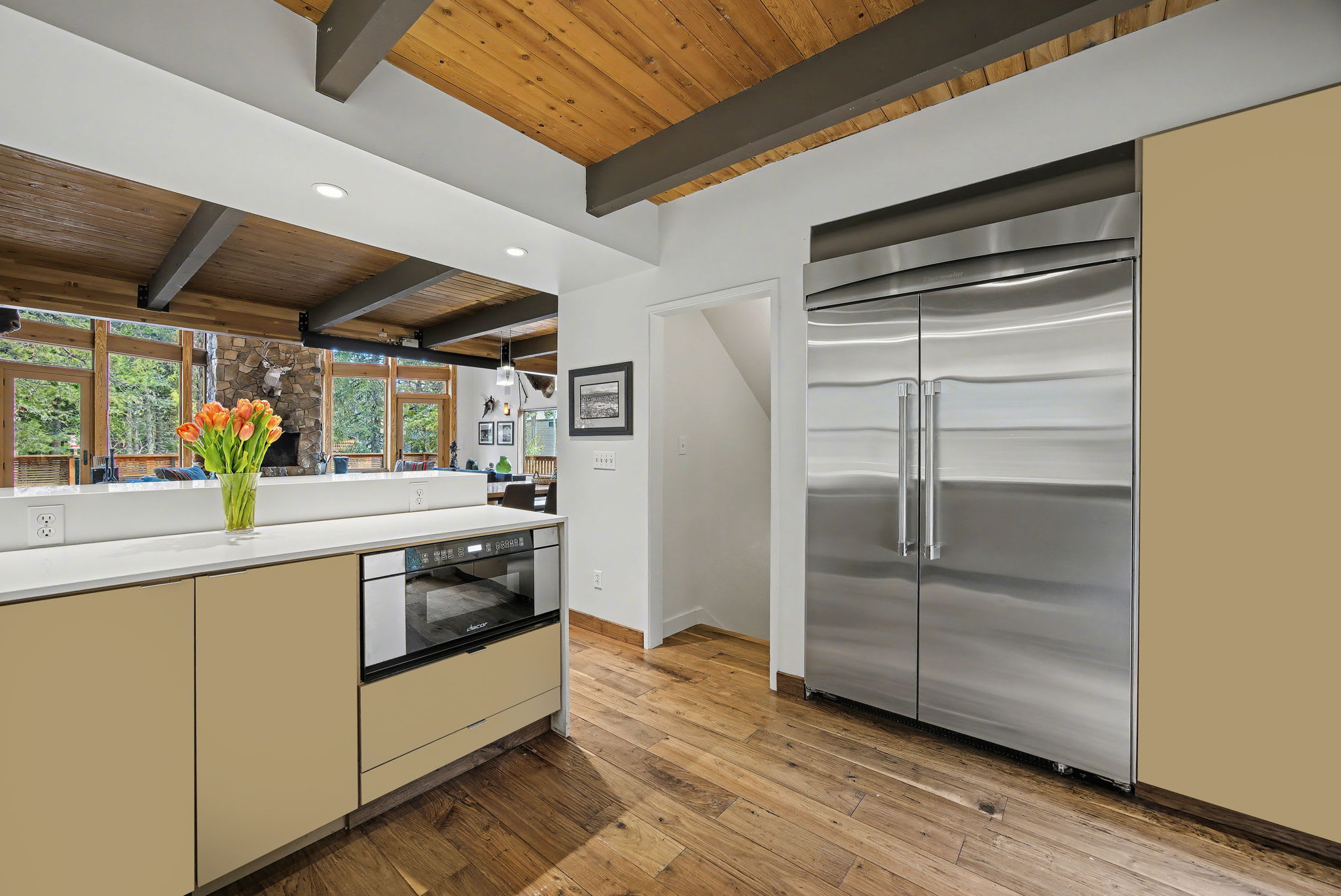This view of the dining area from the end of the long wooden table highlights the open-concept layout leading to the living room. The space features a vaulted wooden ceiling, modern pendant lights, and a built-in bar with a wood-burning stove.