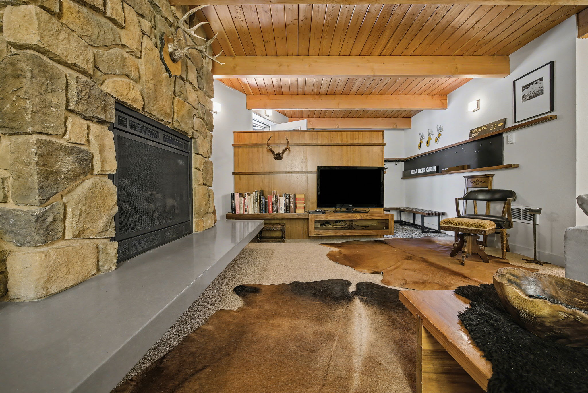 Rustic living room with a large stone fireplace and hearth, a wooden ceiling with exposed beams, and a TV entertainment center with bookshelves, complemented by cowhide rugs on the carpeted floor.