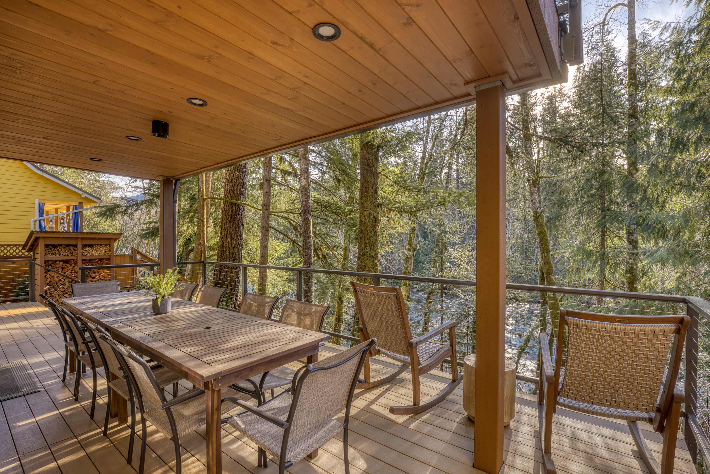 A wide shot of a covered wooden deck with a large outdoor dining table and chairs, offering expansive views of the sun-dappled forest and a glimpse of the river below.