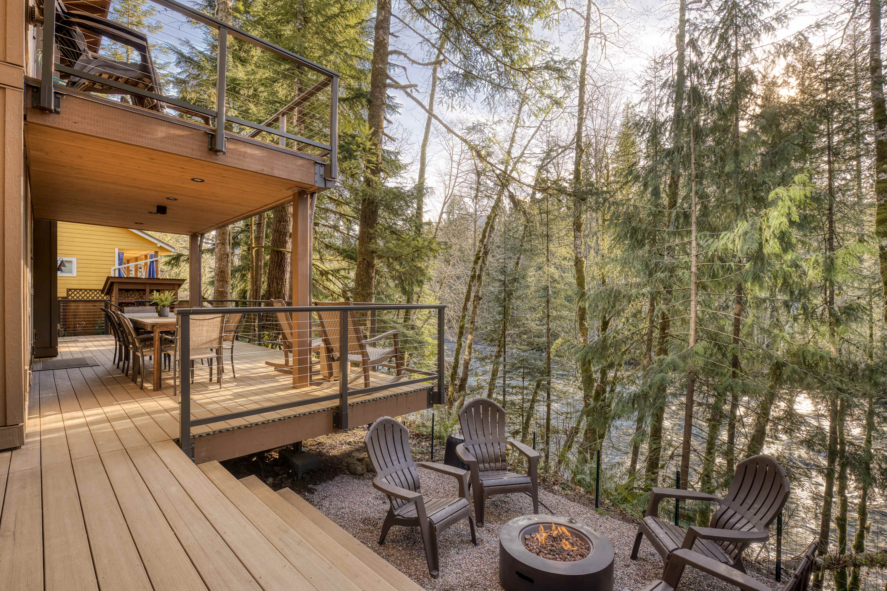A multi-level outdoor space features a wooden deck with stairs leading down to a gravel patio area, furnished with Adirondack chairs and a lit fire pit, all overlooking a river and dense forest.