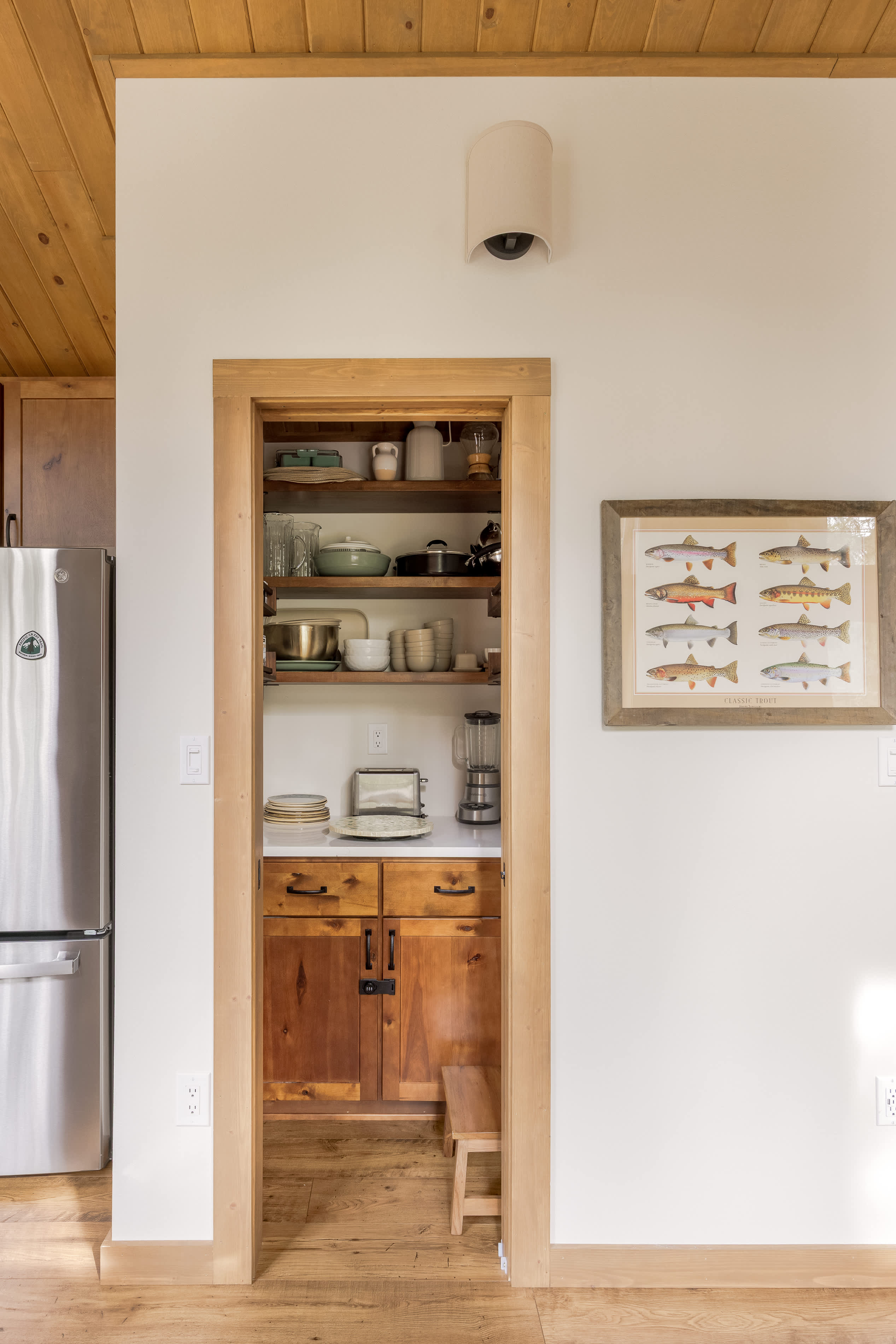 Walk-in pantry area with open wooden shelves stocked with dishes, pots, and a blender, above a lower cabinet with a toaster. A stainless steel refrigerator is visible to the left.