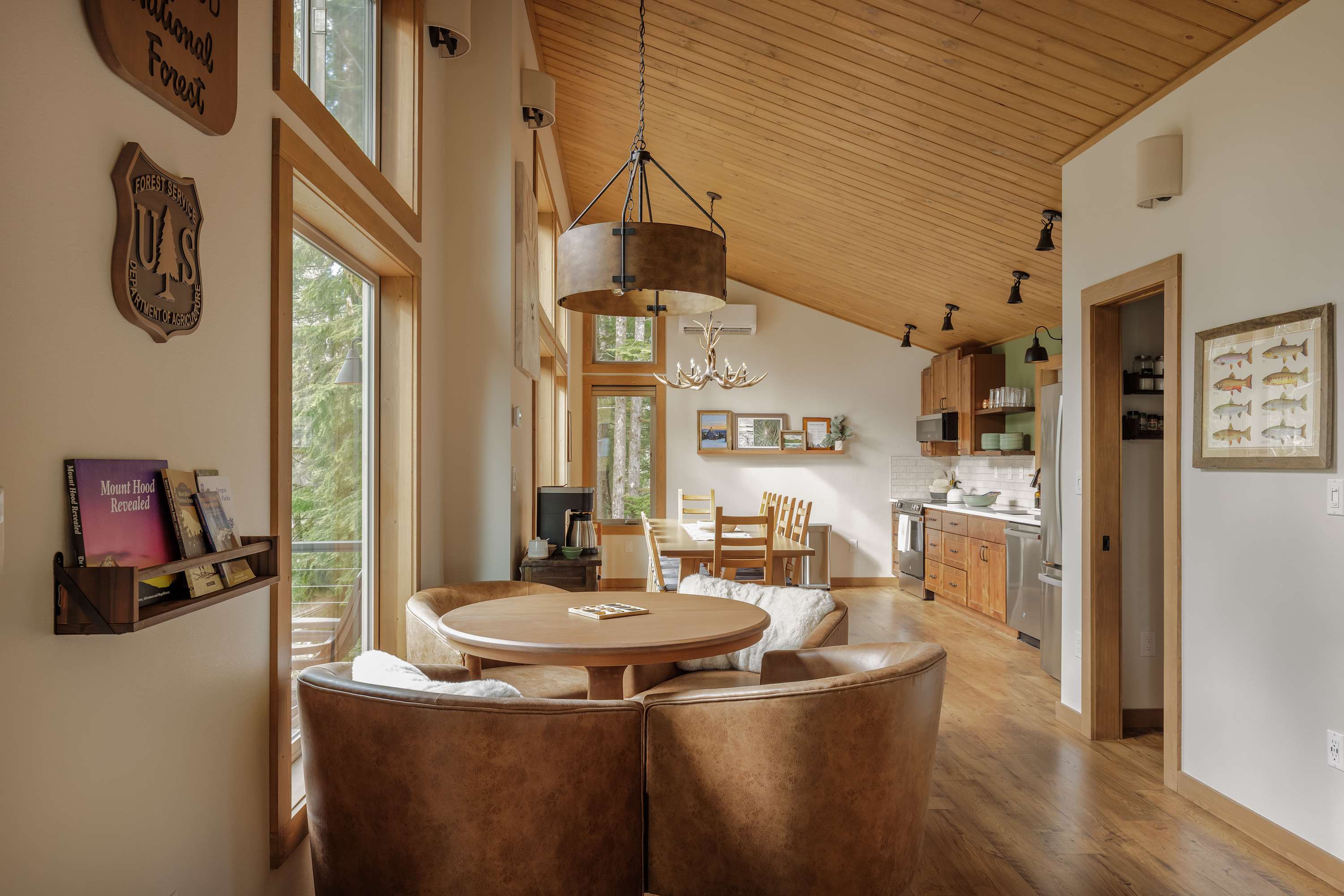 Open-concept dining area with a round wooden table and four brown leather chairs, illuminated by a large circular chandelier. Beyond, a full kitchen with wooden cabinets and stainless steel appliances is visible.