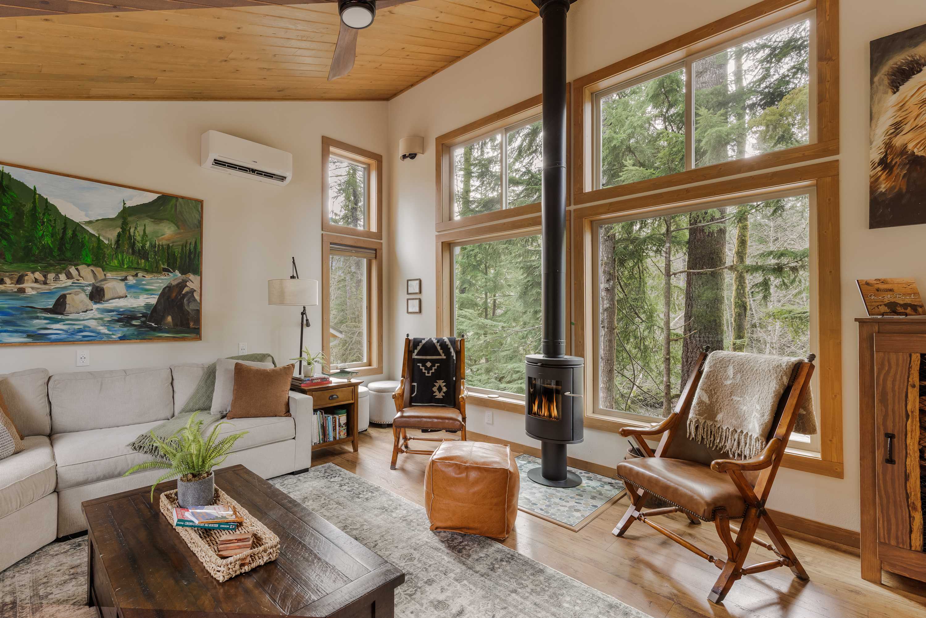 Cozy living room with a light gray sofa, two leather armchairs, a modern black wood-burning stove, and large windows offering views of a lush forest. The room features a wood-paneled ceiling and a large landscape painting.