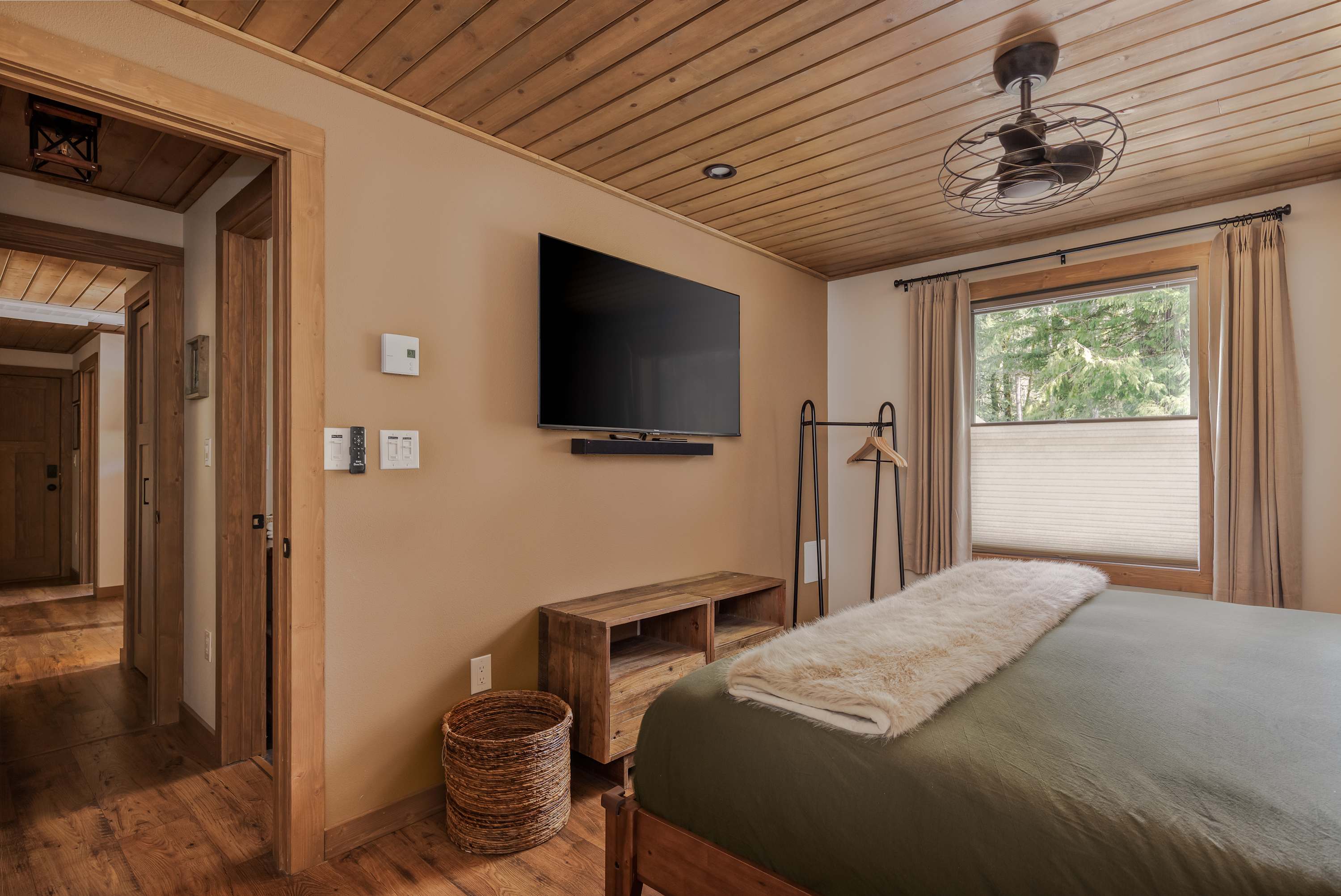 This bedroom view highlights a large window with blinds and curtains, offering a glimpse of the surrounding forest. A simple black clothing rack stands near the window, and a woven basket sits on the hardwood floor, complementing the rustic wooden ceiling.