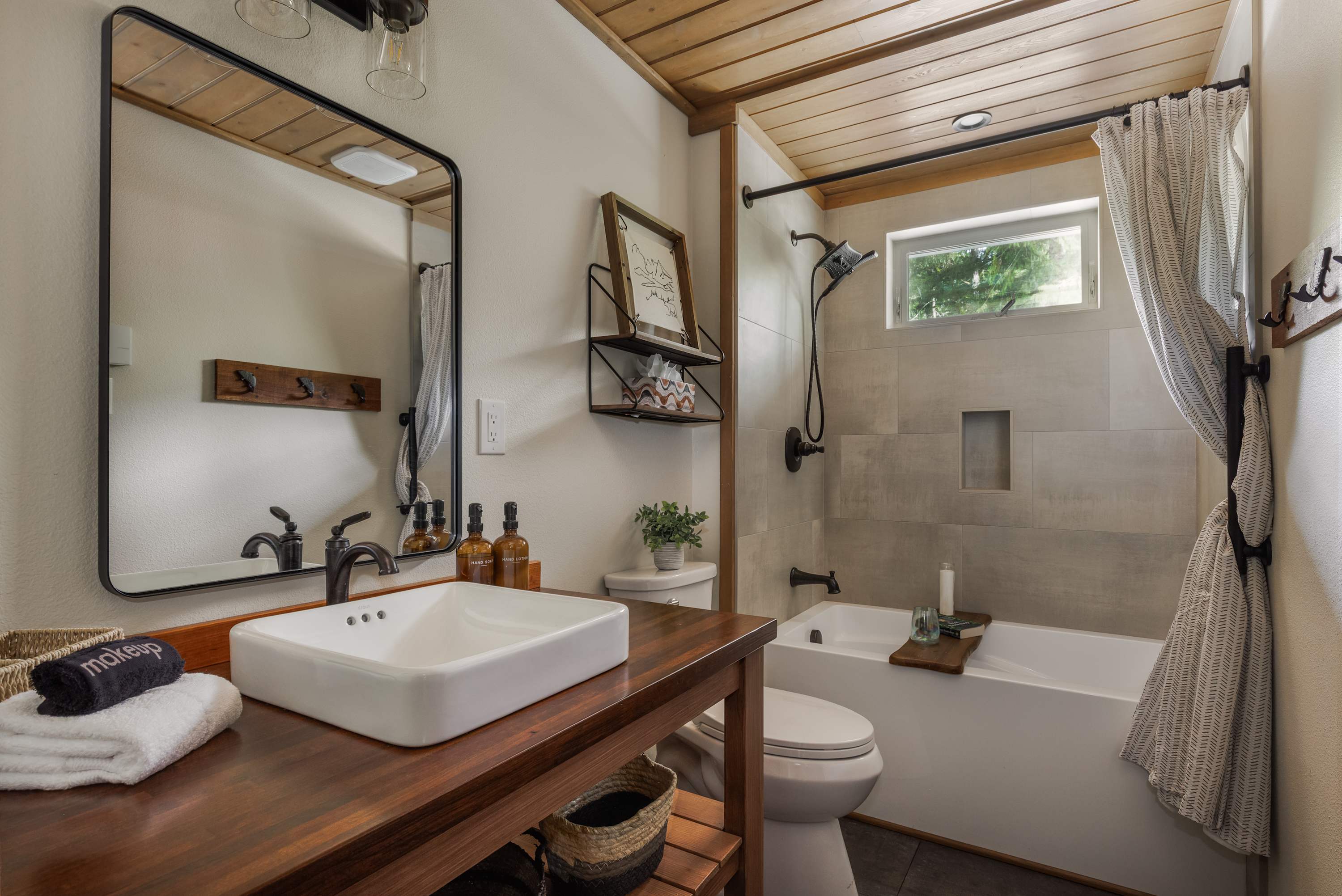 A modern bathroom features a rectangular vessel sink on a dark wood vanity with an industrial-style mirror. The shower/tub combo has light gray subway tiles, a niche, and a window, with a patterned shower curtain adding a touch of texture.