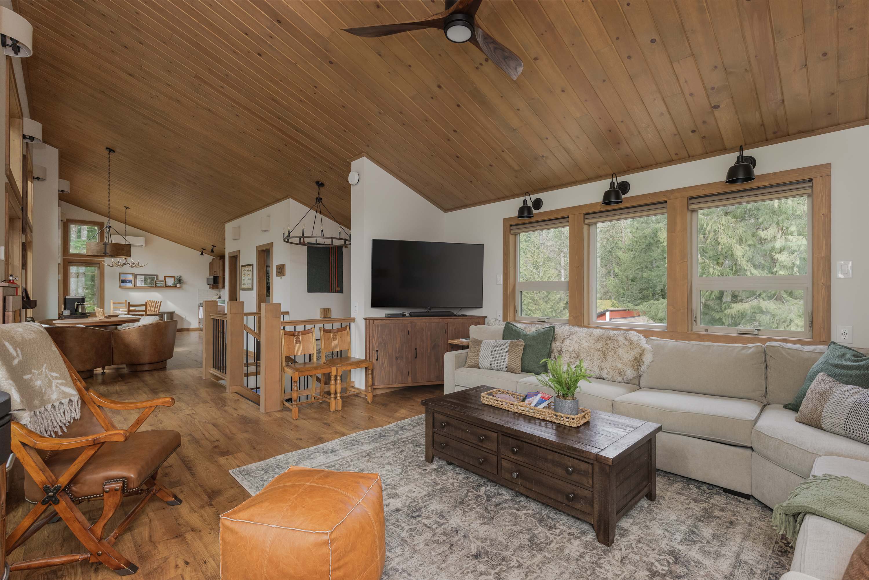 An open-concept living room with a large flat-screen TV on a wooden console, a sectional sofa, and views into the dining area. The room features hardwood floors, a vaulted wood ceiling, and ample natural light.