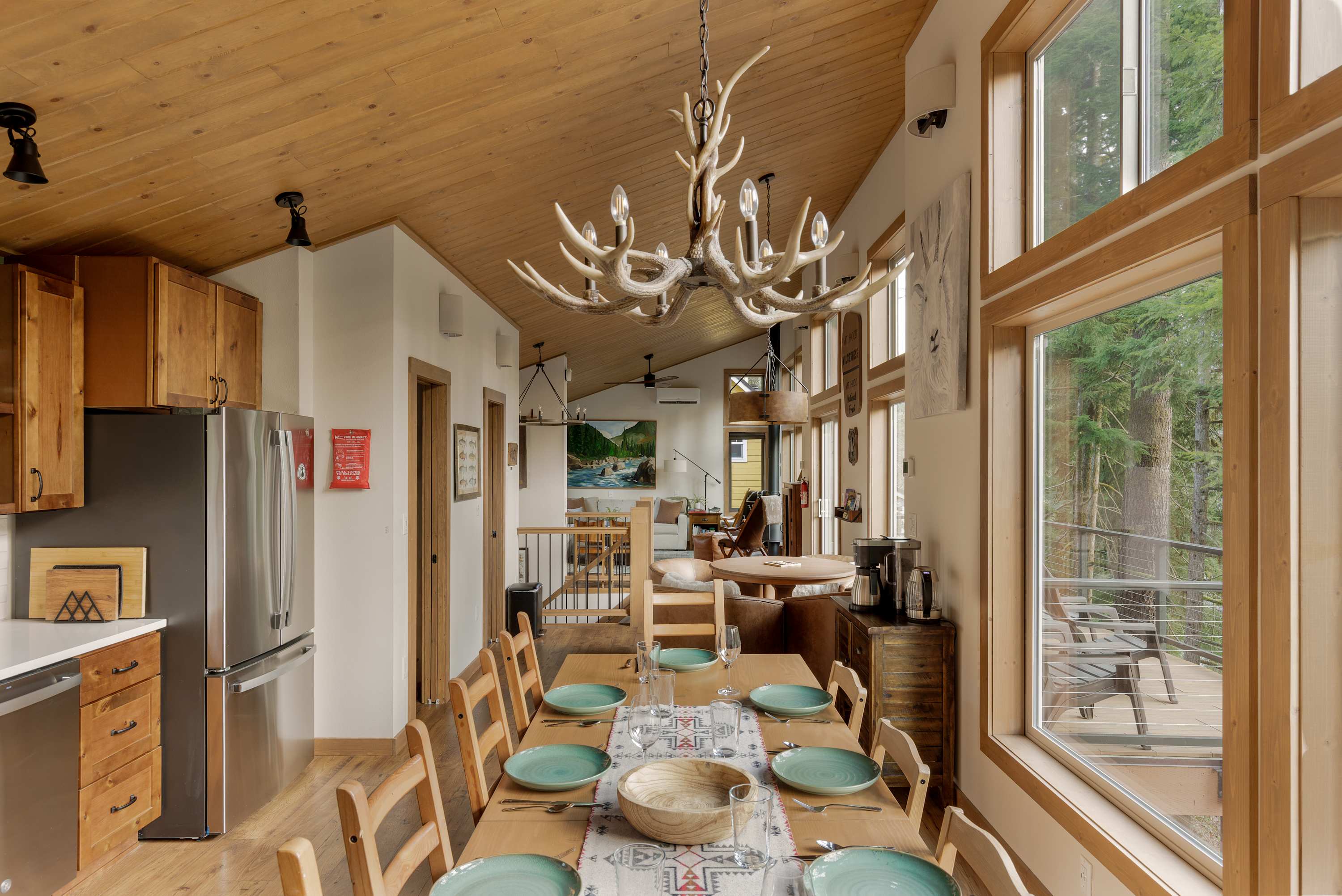 Spacious dining room featuring a long wooden table set with green plates, illuminated by an antler-style chandelier. The adjacent kitchen with wooden cabinetry and stainless steel appliances is visible.