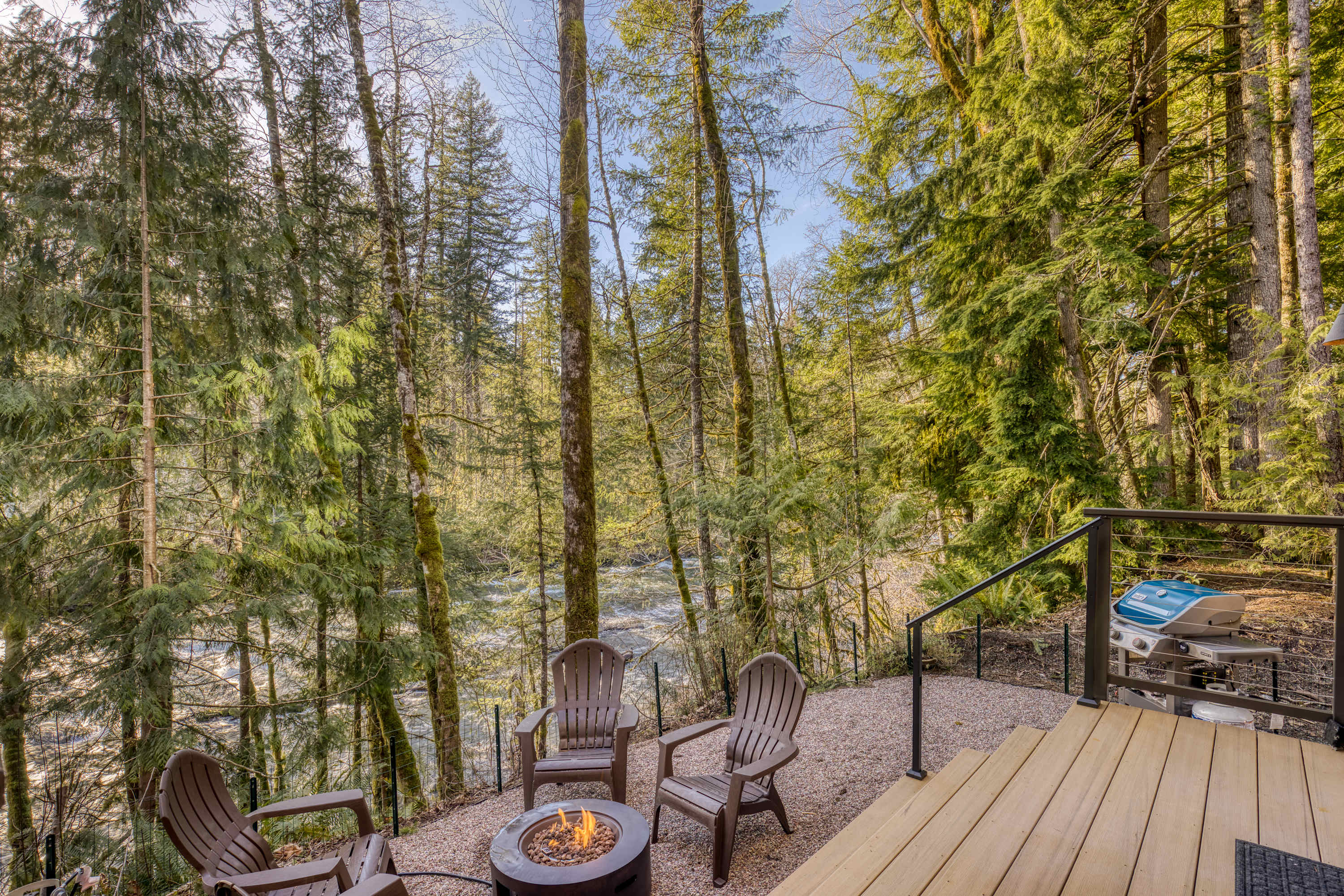 A cozy gravel patio area features a circular fire pit surrounded by dark Adirondack chairs, with a BBQ grill visible on the adjacent wooden deck, all set against a backdrop of a river and dense forest.