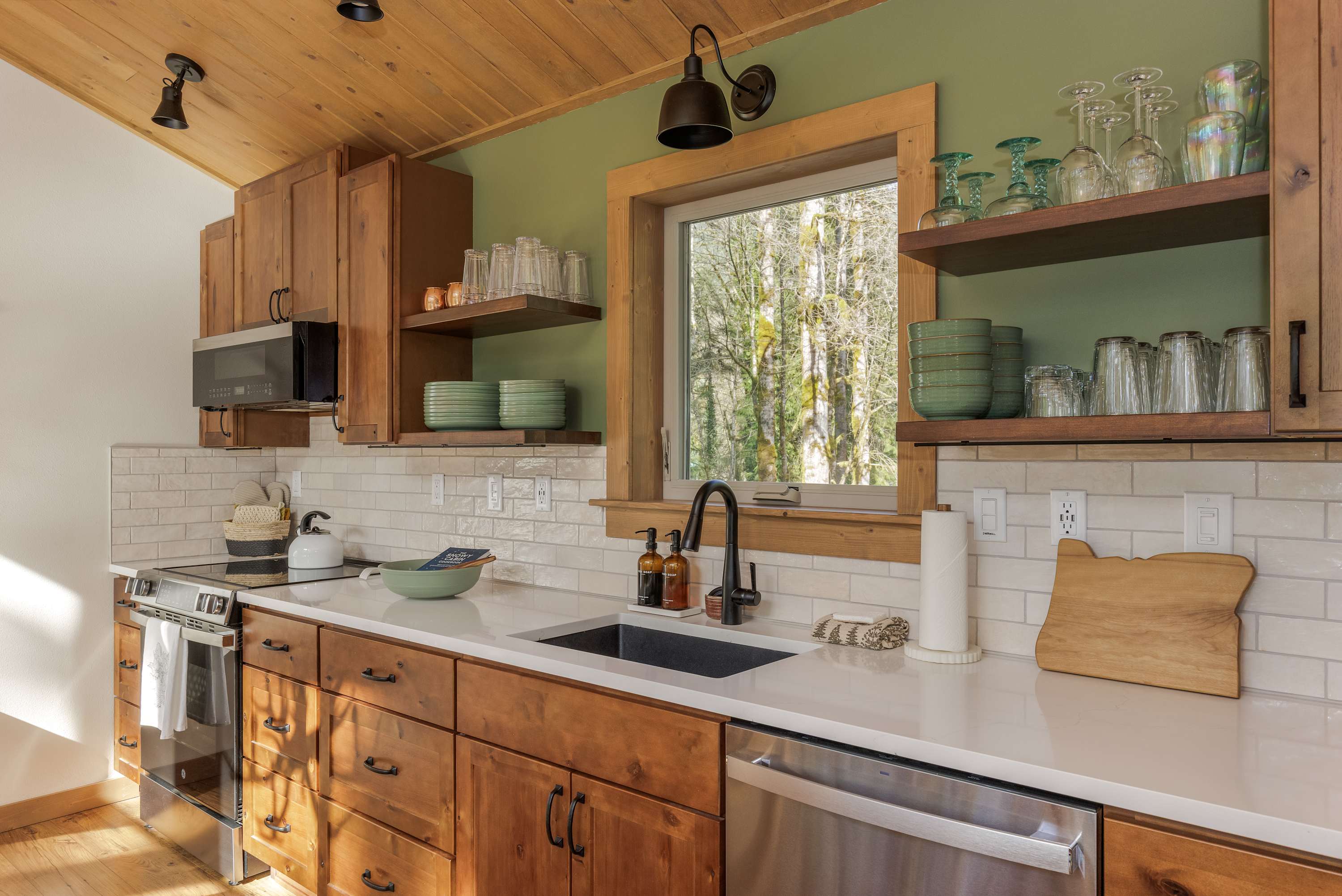 Modern kitchen with light wood cabinetry, white subway tile backsplash, and white countertops. Open shelving displays dishware, and a window offers a view of the surrounding trees.