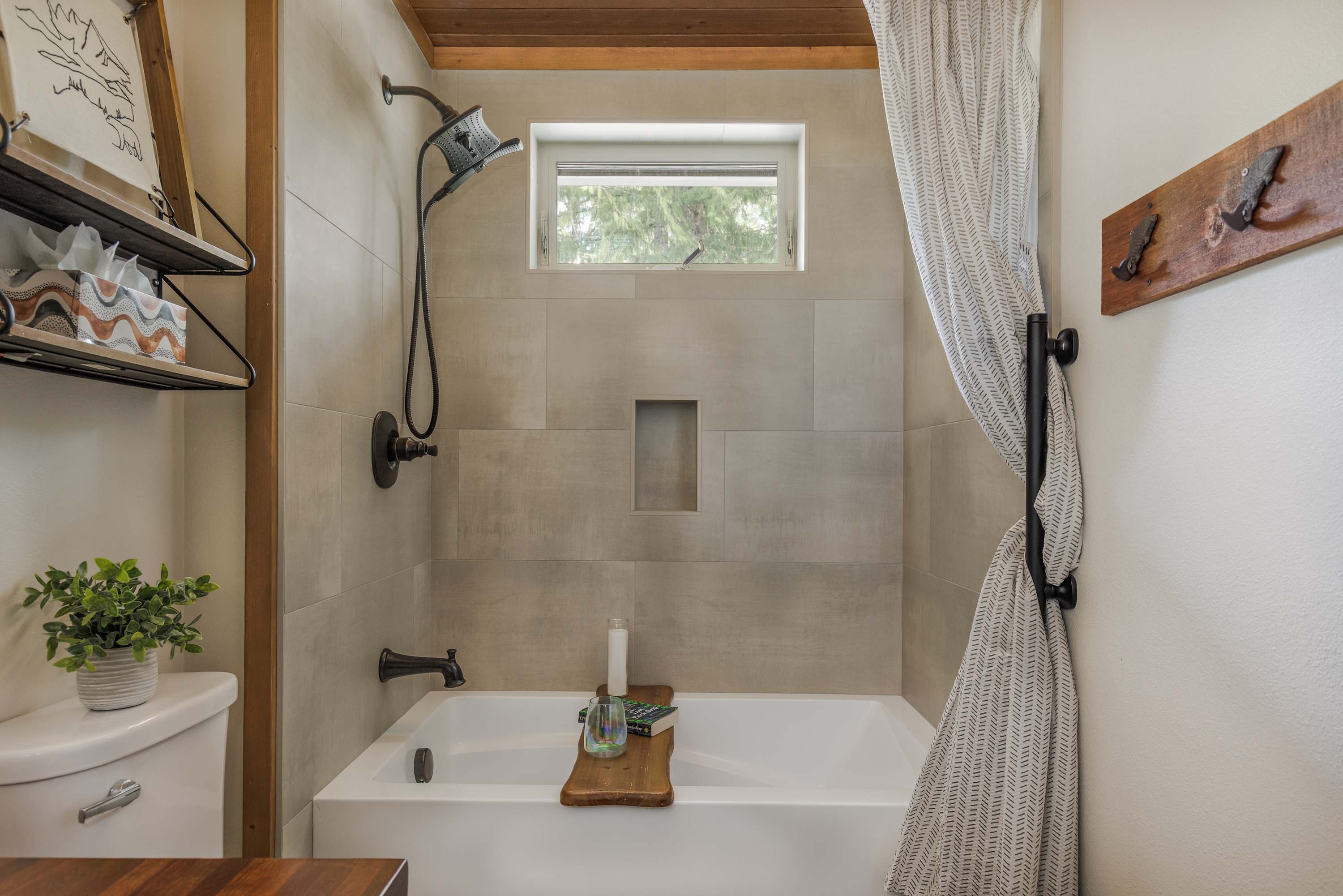 A close-up of the bathtub area in the bathroom, featuring light gray subway tiles, a built-in shower niche, and a small window above the tub. A dark bronze shower head and faucet complement the modern, clean design, with a wooden bath caddy resting on the tub.