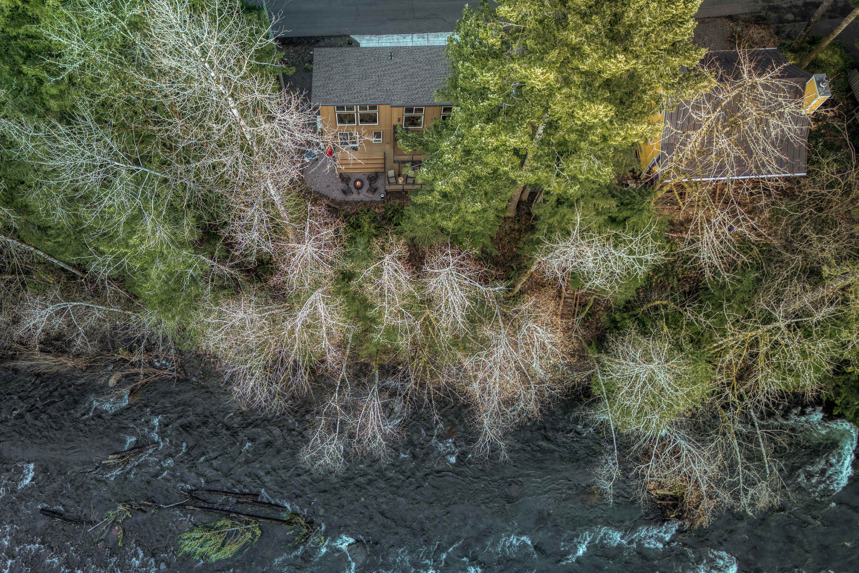 A high aerial view captures a house nestled among a dense forest of evergreen and bare deciduous trees, with a winding, turbulent river flowing alongside the property.