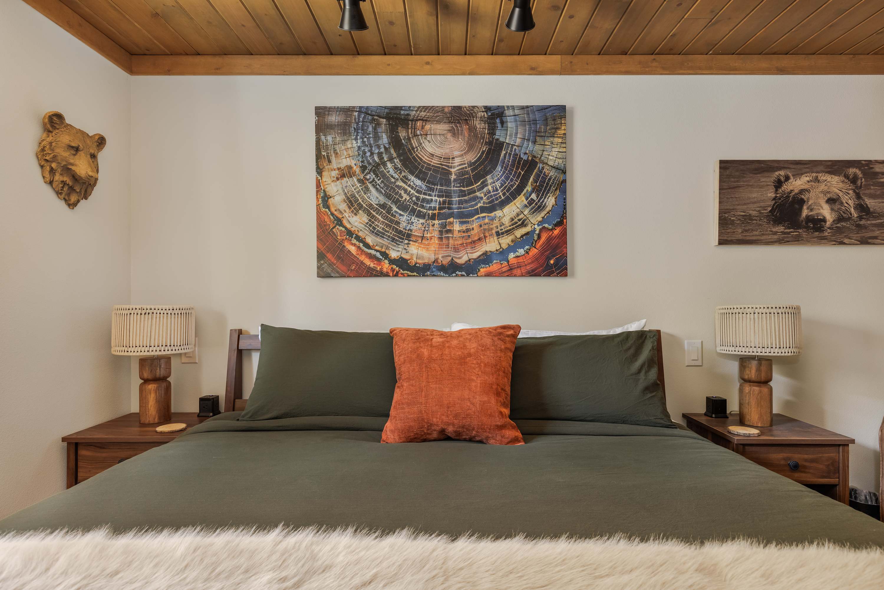 A close-up of the king bed in the bedroom, featuring dark green bedding, an orange accent pillow, and a faux fur throw. The bed is framed by wooden nightstands with woven lamps, and the wall above is adorned with nature-inspired artwork, including a large abstract tree ring print.