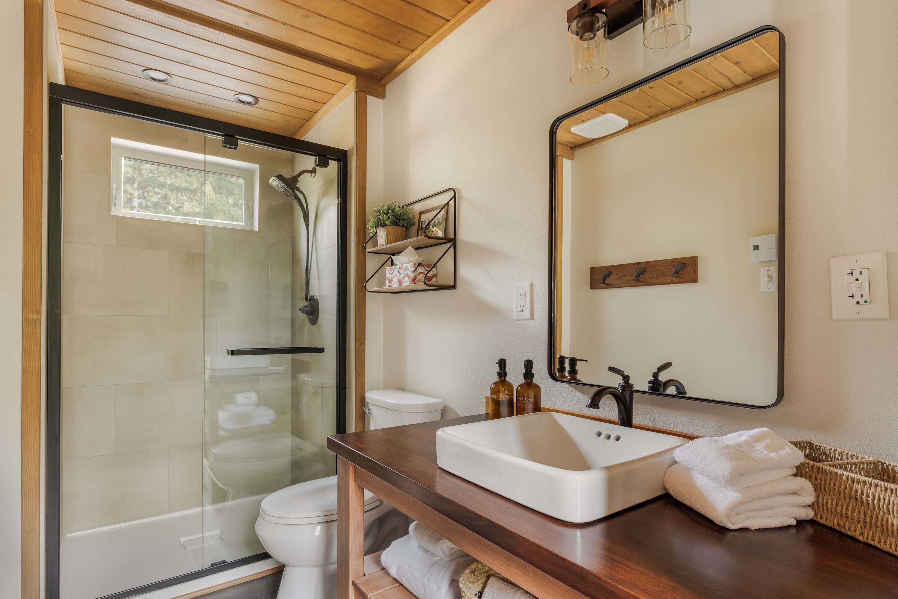 A modern bathroom with a walk-in shower featuring a glass enclosure and a small window. A dark wood vanity with a white vessel sink, a large black-framed mirror, and industrial-style lighting completes the space.