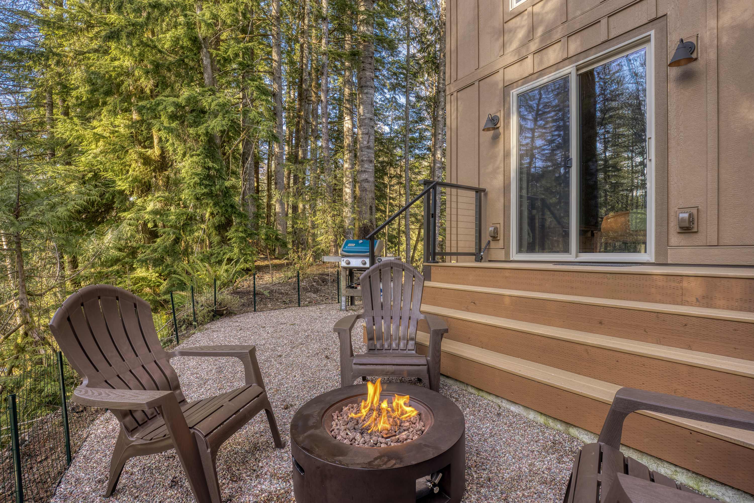 An inviting outdoor patio area features a circular fire pit with active flames, surrounded by three brown Adirondack-style chairs on a gravel surface. A wooden deck with stairs leads up to the house, which is nestled against a dense forest backdrop.
