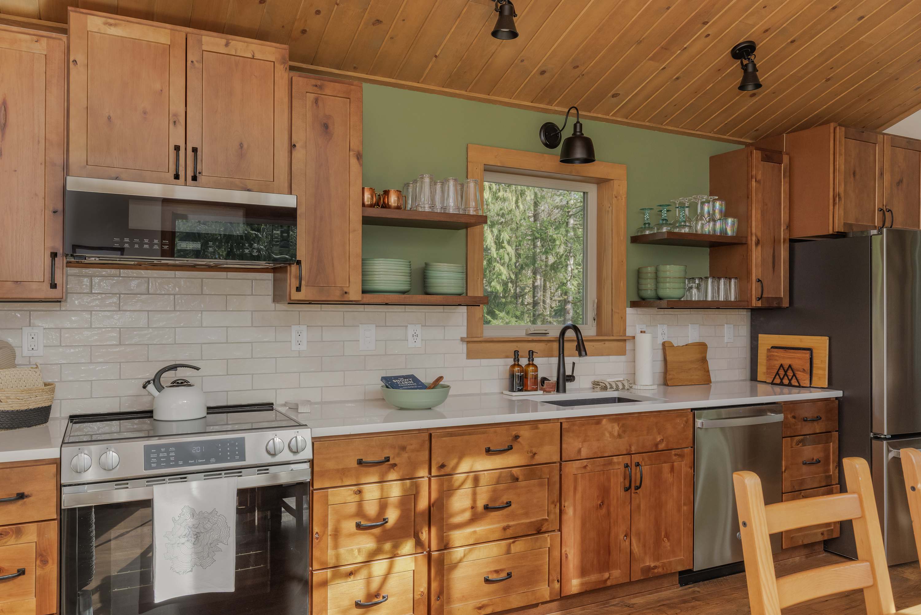 Well-equipped kitchen featuring light wood cabinets, white countertops, and a white subway tile backsplash. Stainless steel appliances include a range, microwave, and refrigerator, with a window overlooking the forest.