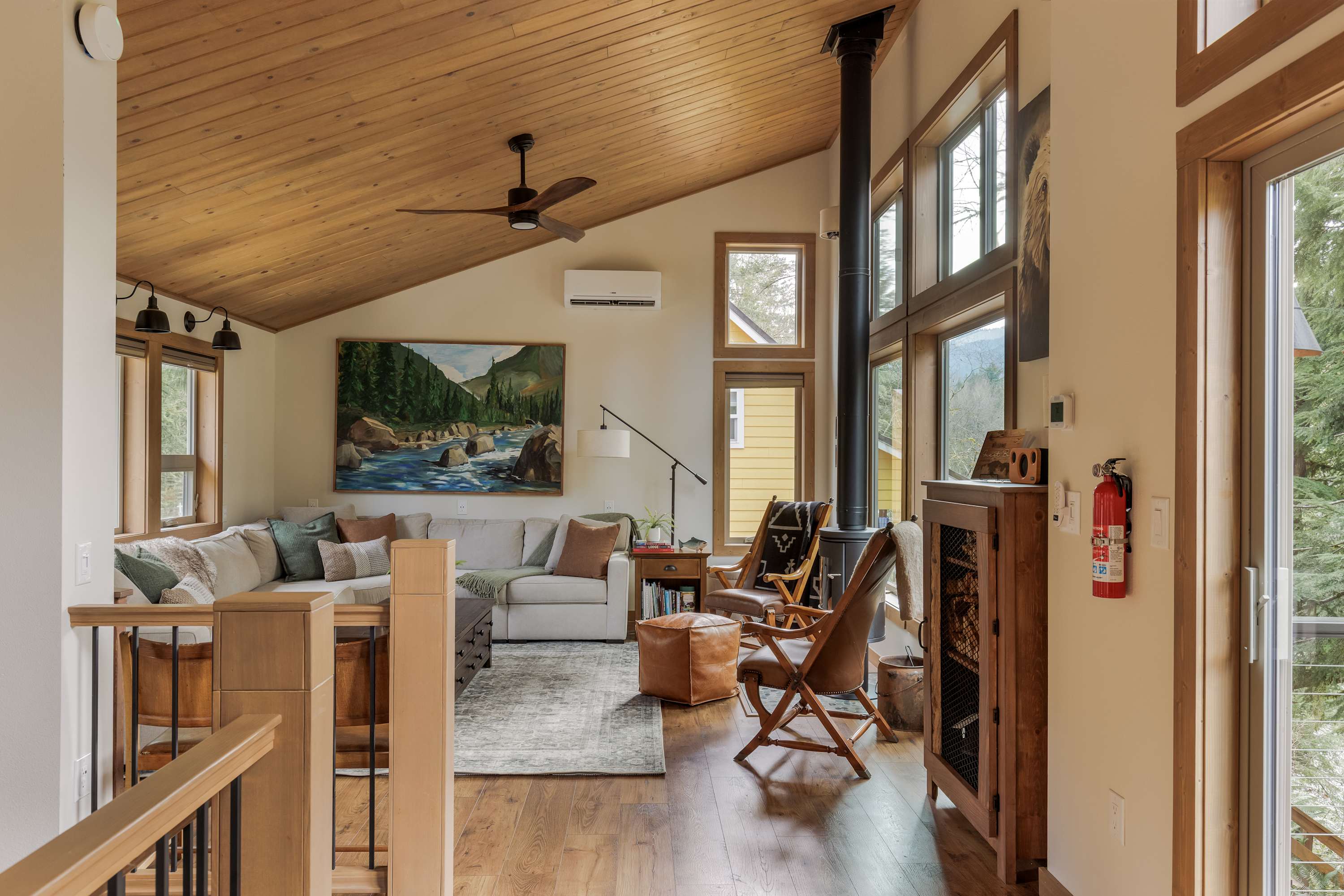 A spacious living room with a vaulted wood-paneled ceiling and hardwood floors, featuring a large sectional sofa and a modern wood-burning stove. Large windows offer views of the surrounding trees.