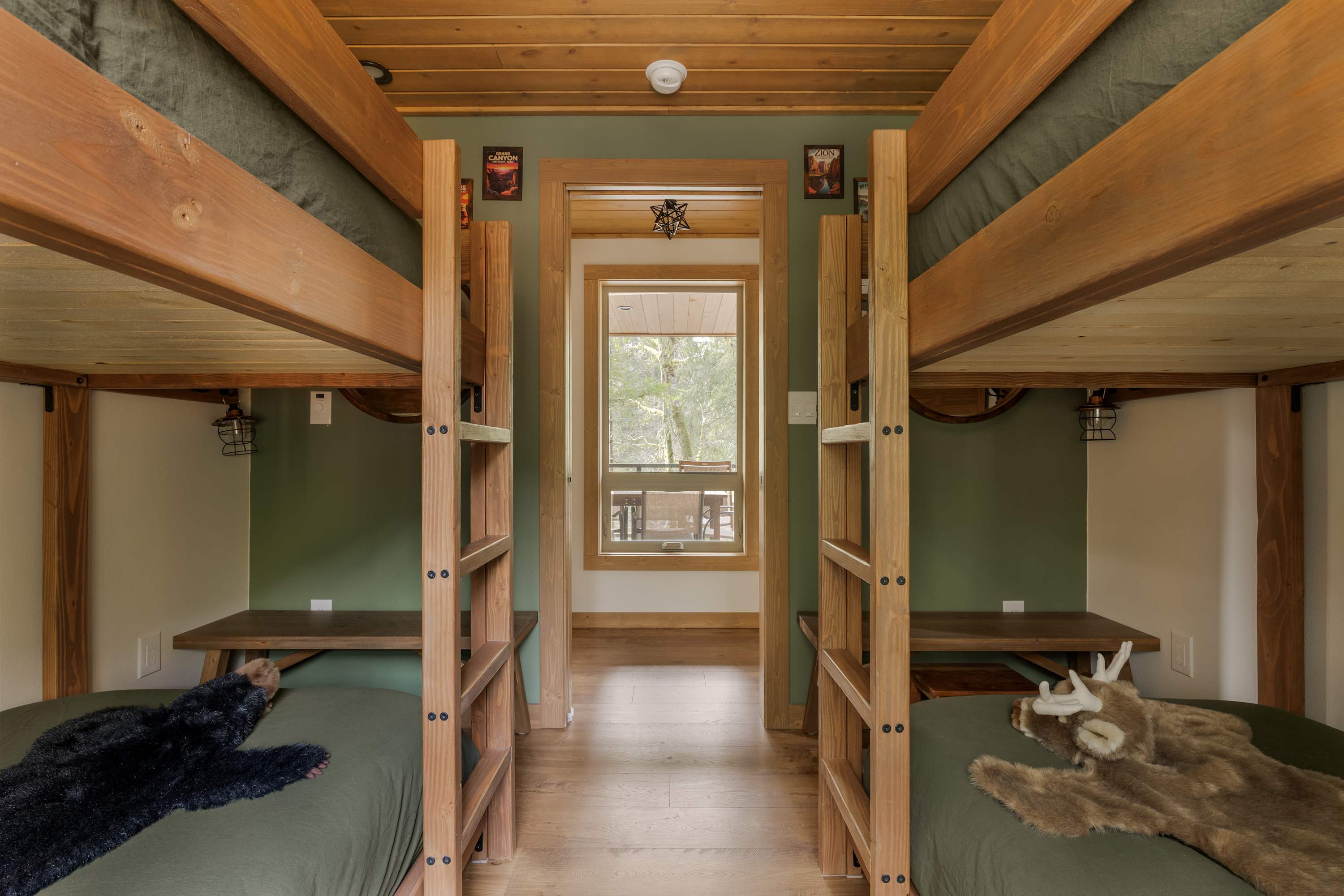 A bunk room with two sets of sturdy wooden bunk beds, each with dark green bedding and animal-themed plush toys. A window at the far end offers a view of trees, and the room has a wooden plank ceiling.