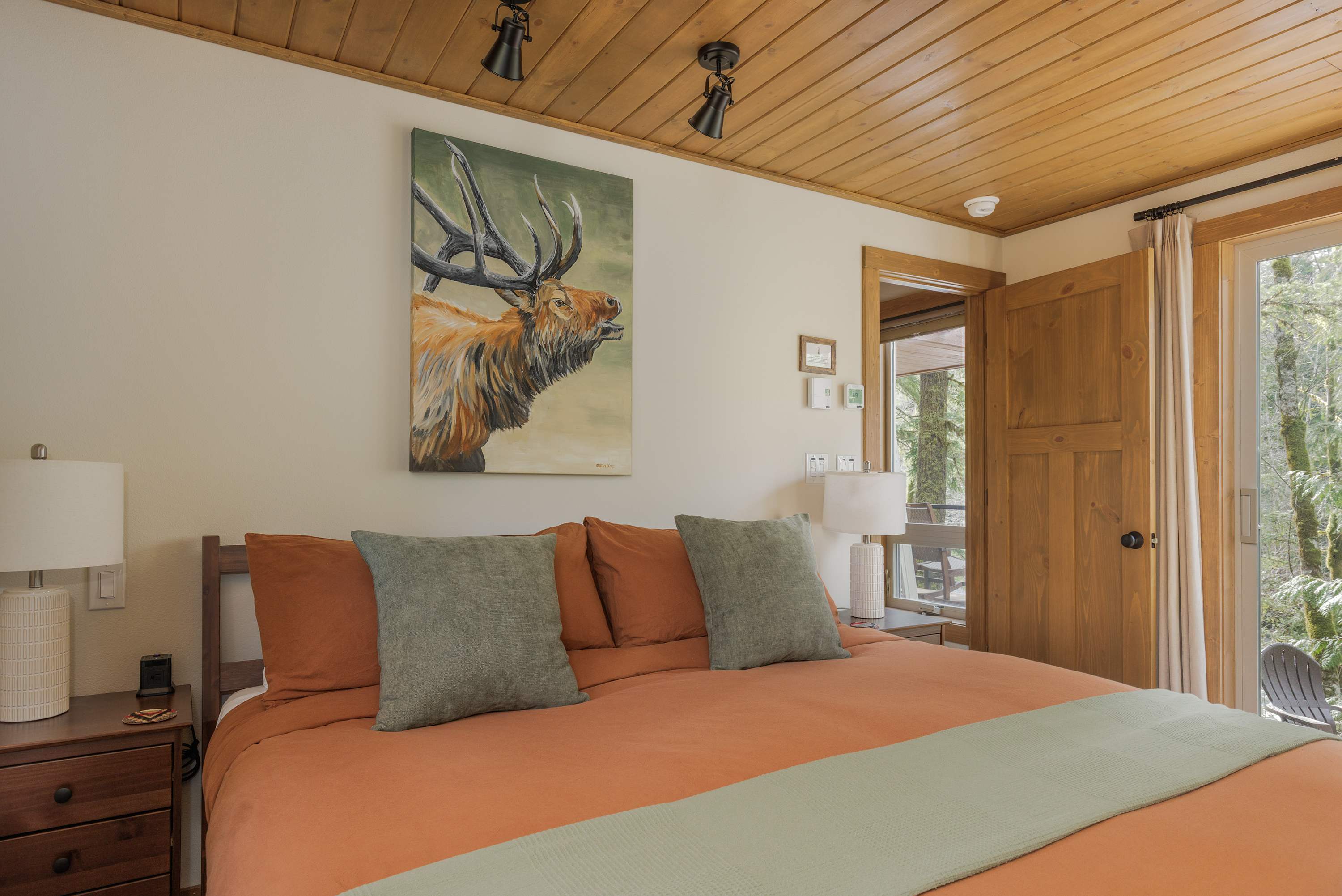 View of a cozy bedroom featuring a large bed with orange bedding, green pillows, and wooden nightstands with lamps. An elk painting adorns the wall, and a wooden door leads to an outdoor space with trees visible.