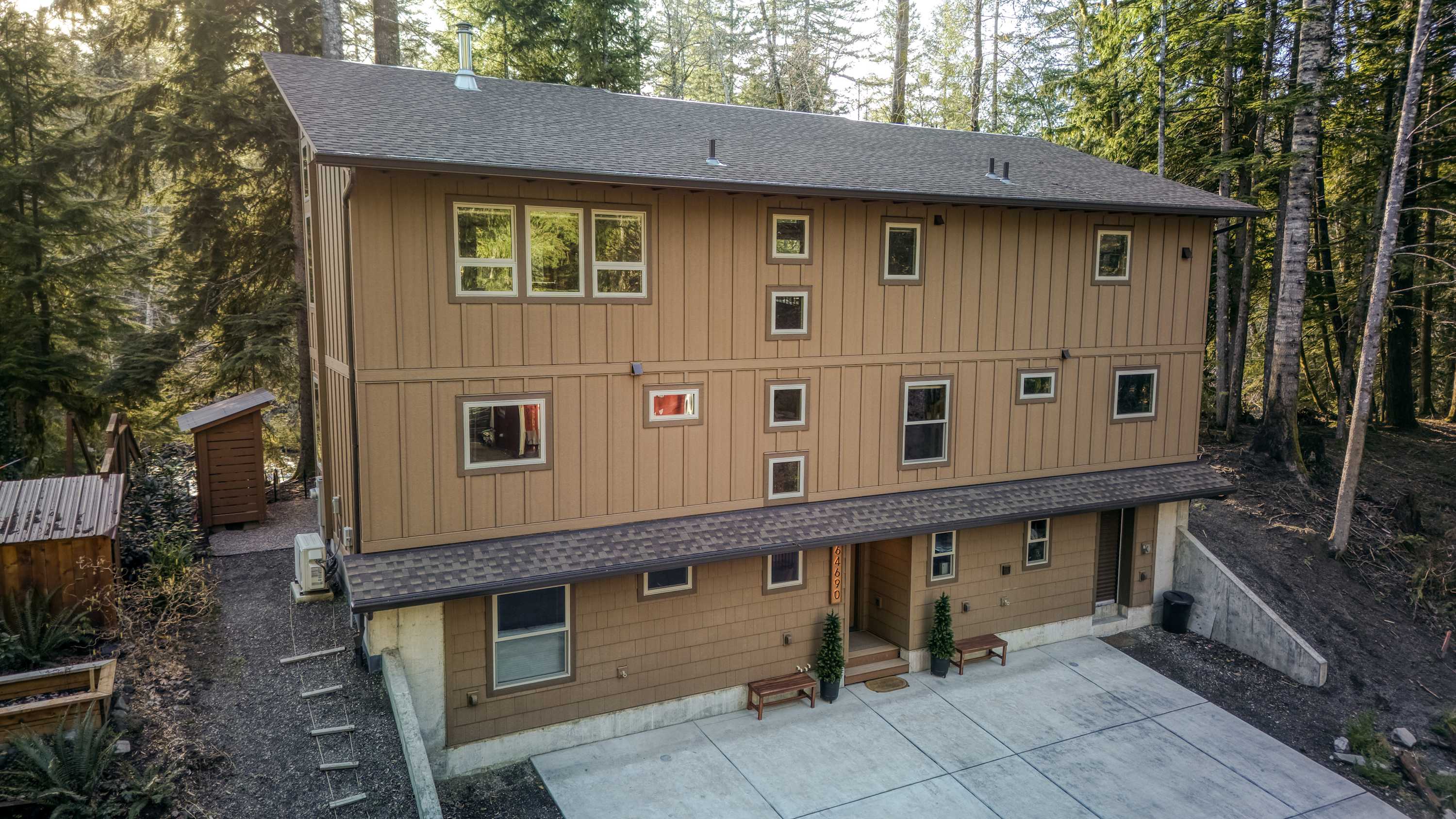 Aerial view of a large, three-story house with brown siding and a dark shingled roof, nestled among tall evergreen trees. A concrete driveway leads to the front entrance, with benches and potted plants flanking the door.
