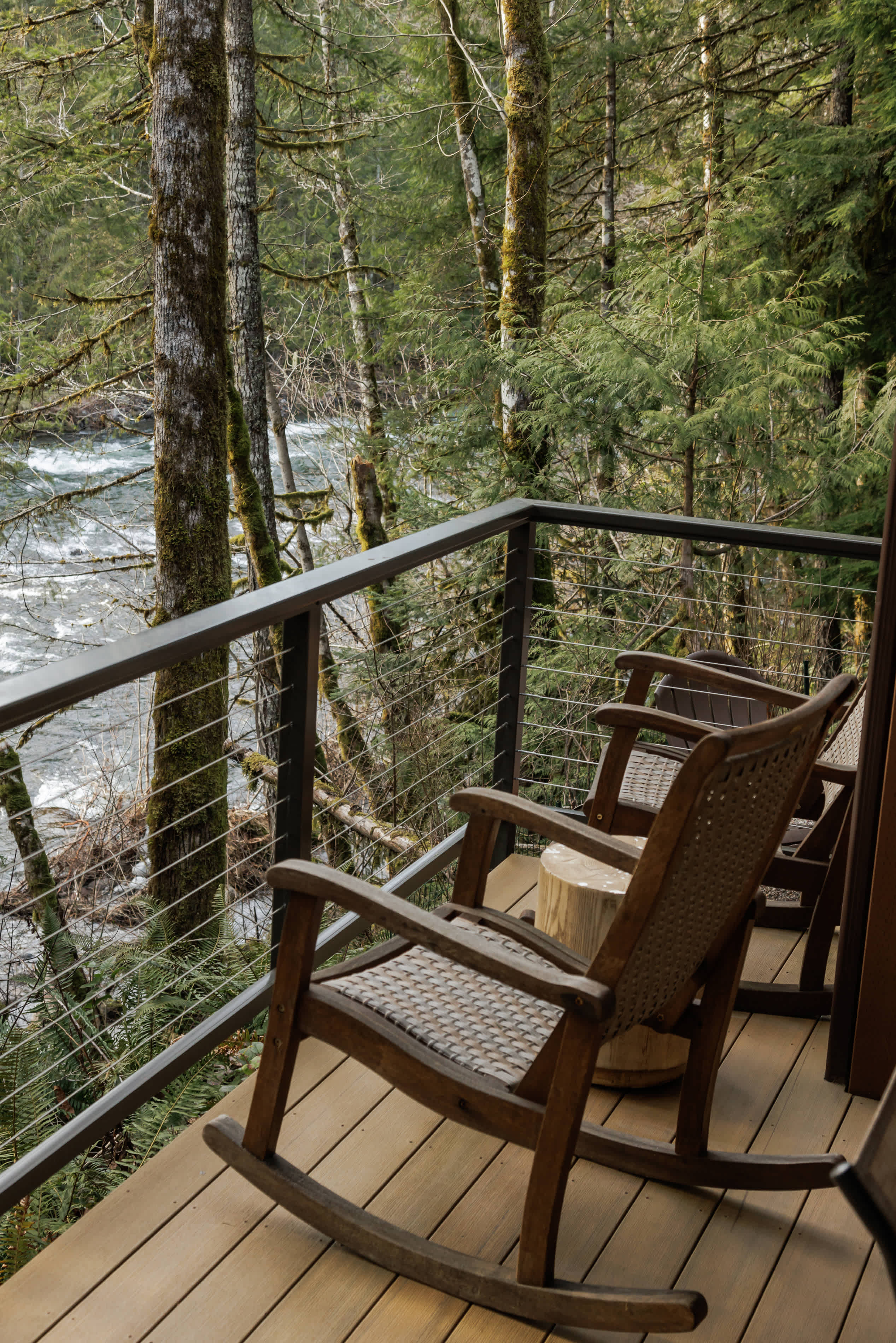 A close-up view of a wooden balcony features two dark rocking chairs with woven seats and a rustic wooden side table, all overlooking a rushing river and dense forest through cable railings.