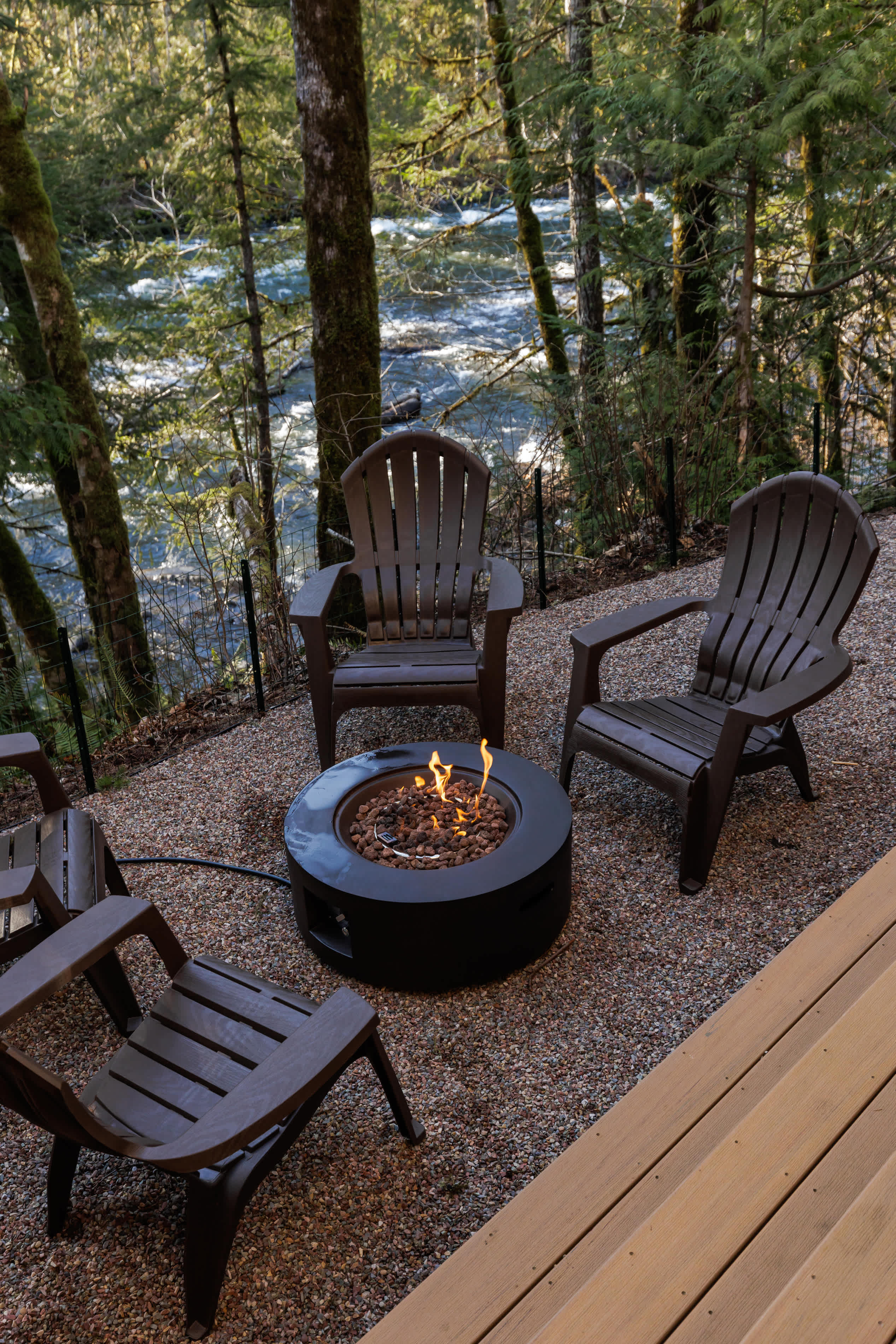 A close-up view of a lit circular fire pit on a gravel patio, surrounded by dark Adirondack chairs, with the rushing river and forest visible in the background.