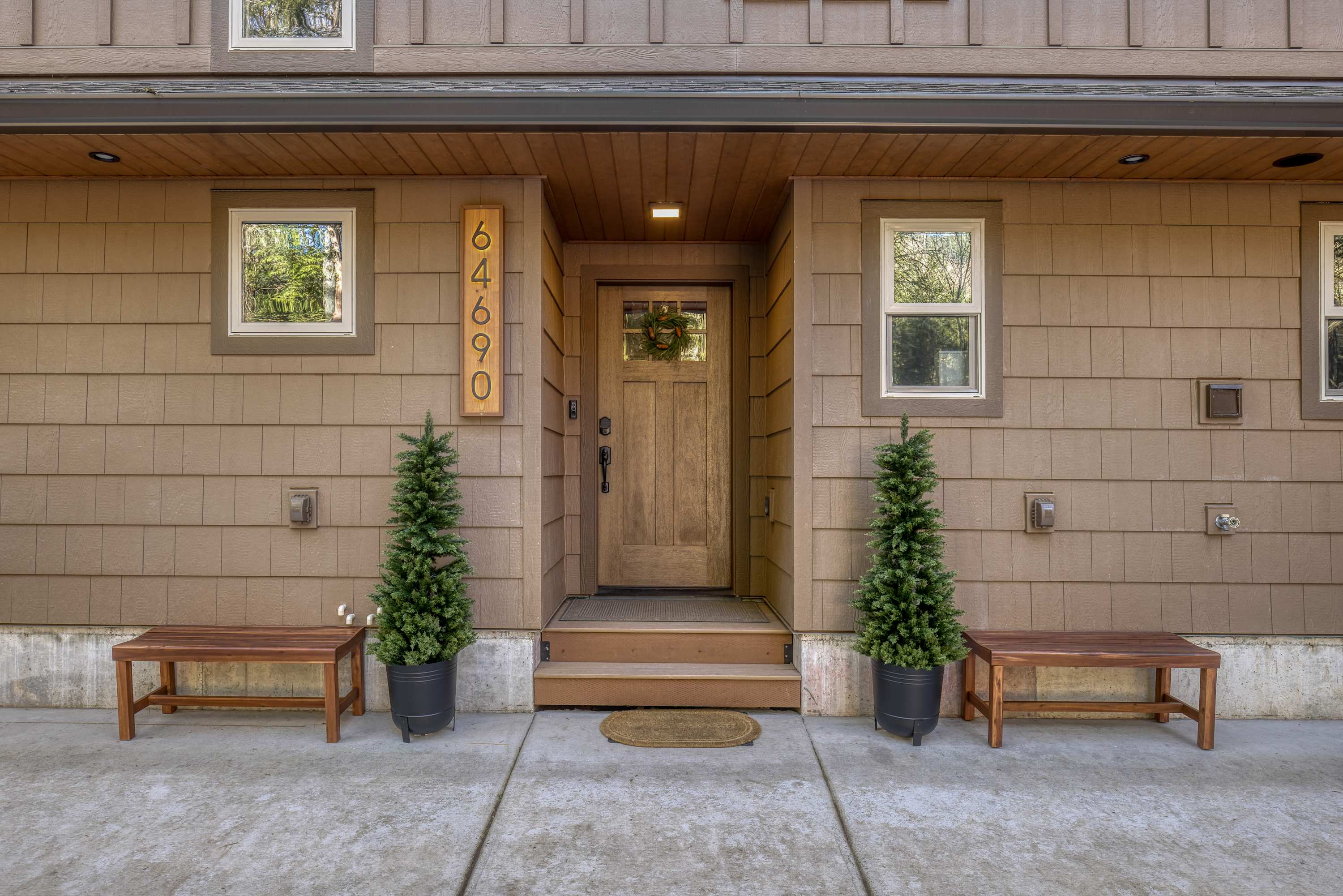 The front entrance of the house, featuring a wooden door with a wreath, flanked by two benches and potted evergreen trees. The exterior is clad in brown shingle siding with white trim windows.