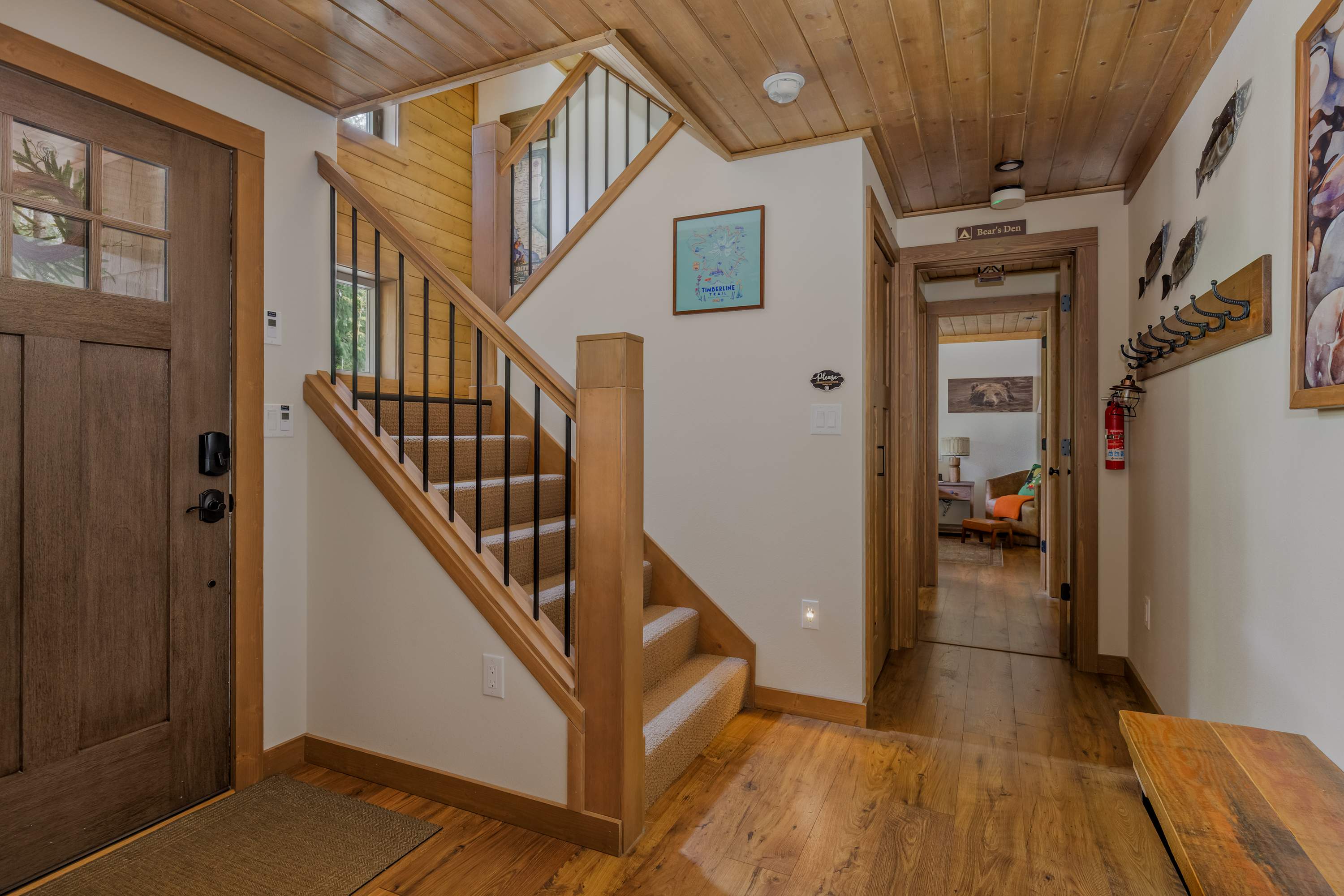 A welcoming foyer with hardwood floors and a wooden staircase leading upstairs, featuring black metal balusters. A wooden door is visible on the left, and a hallway extends to the right.