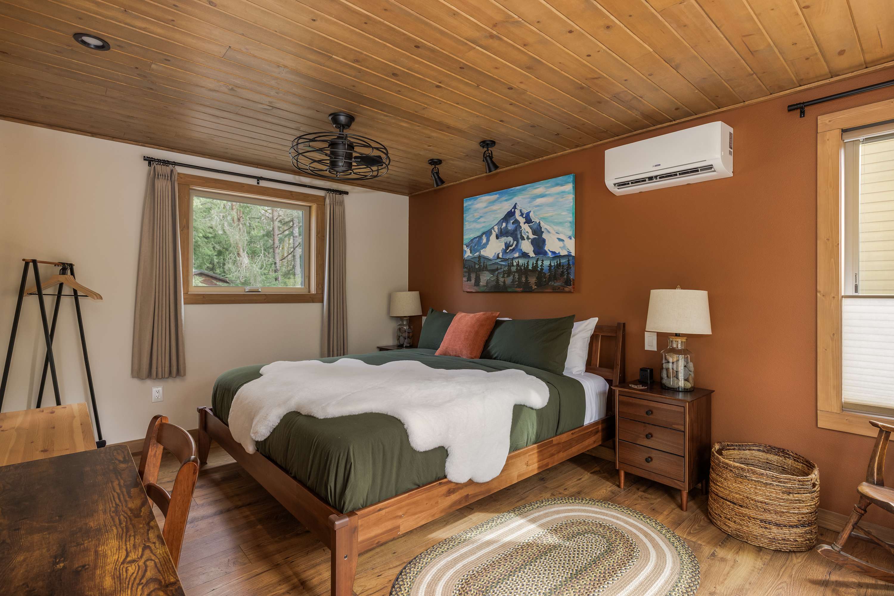 A warm bedroom with a rust-colored accent wall and a wooden plank ceiling. A large bed with dark green bedding and a white faux fur throw is flanked by wooden nightstands with lamps. A mountain landscape painting hangs above the bed.