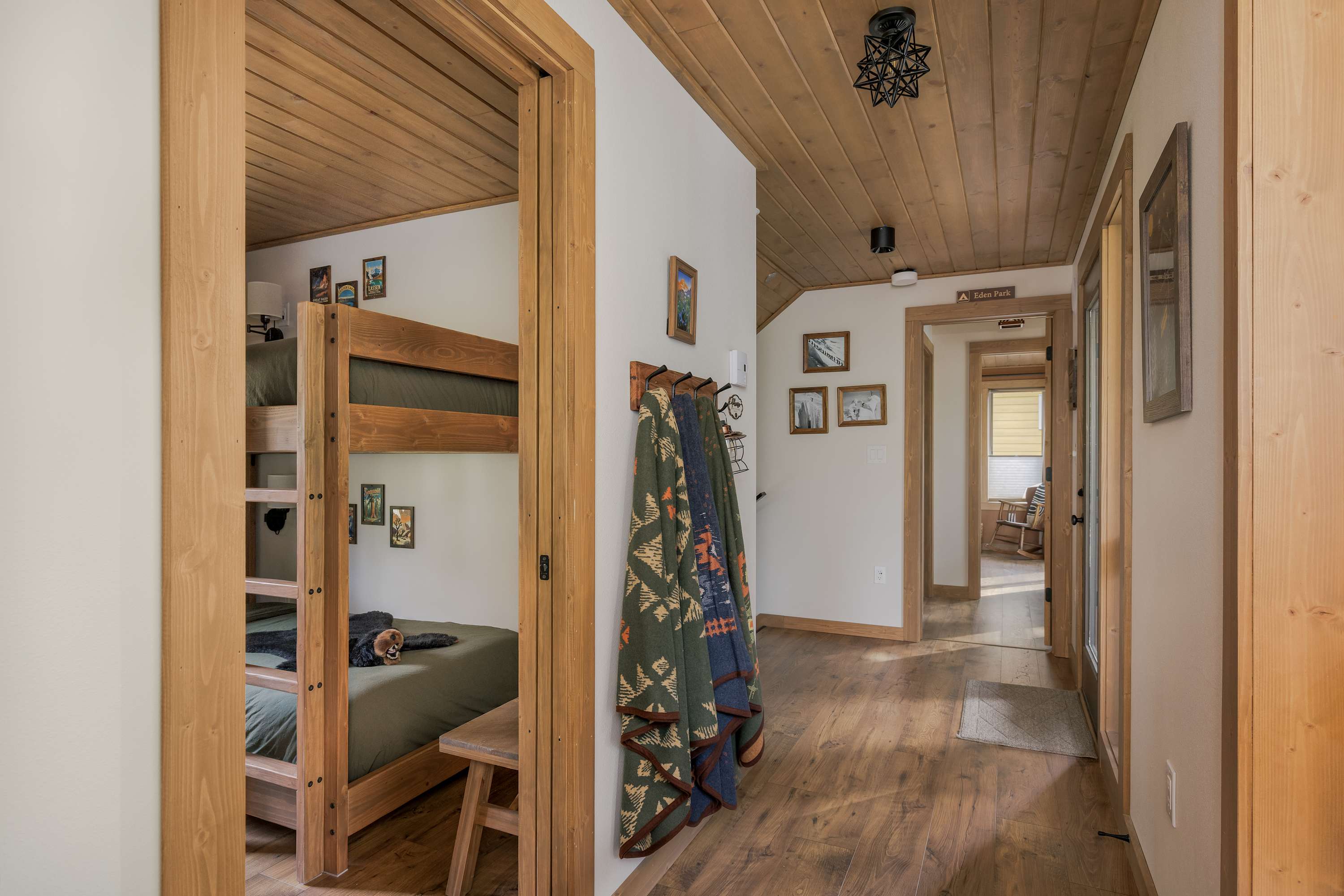A rustic hallway with wooden plank ceilings and hardwood floors, featuring a coat rack with patterned blankets. Doorways lead to other rooms, including a bunk room visible on the left.
