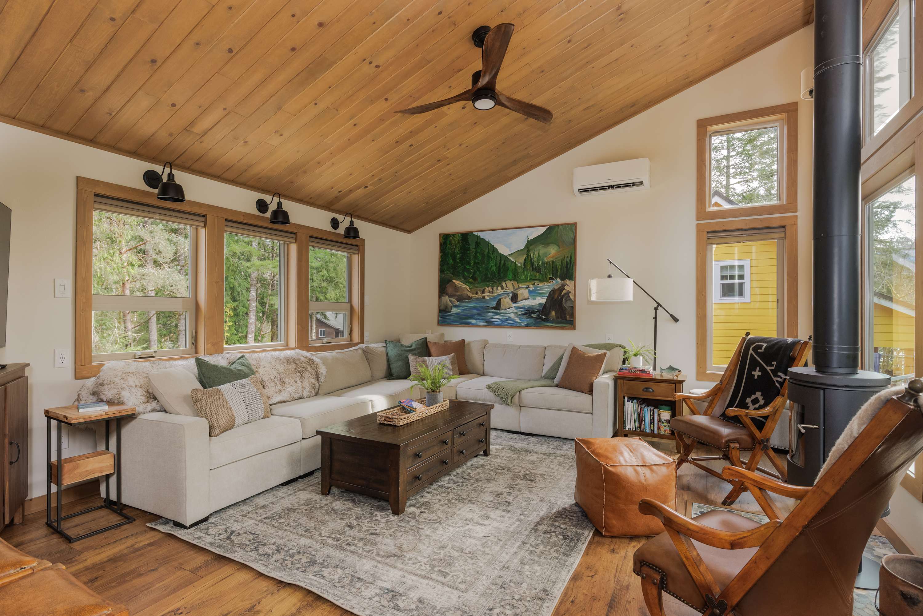 A wide view of the living room showcasing a large light-colored sectional sofa, a wooden coffee table, and a patterned rug on hardwood floors. The room has a vaulted wood ceiling and multiple windows.