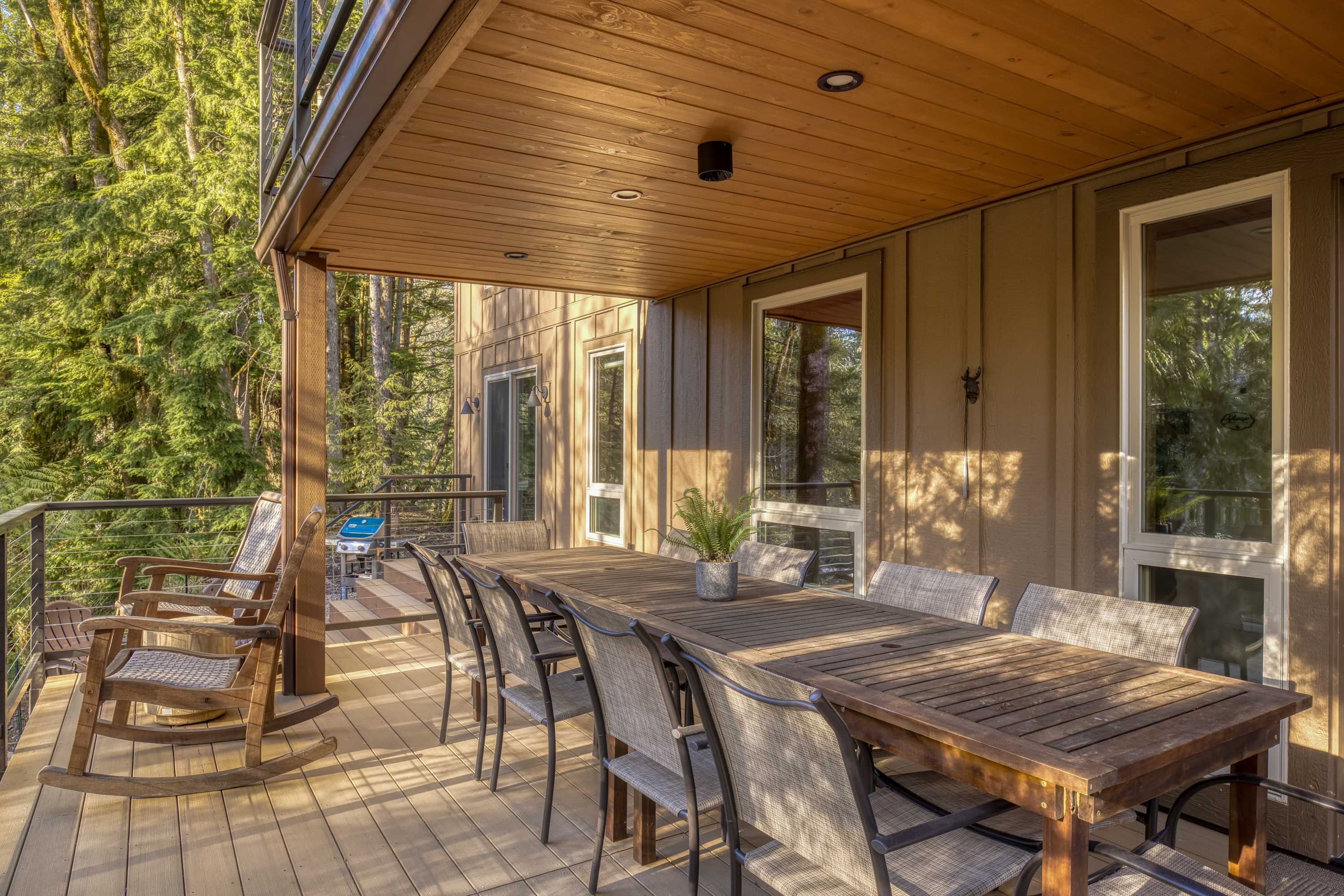 A spacious covered wooden deck features a long wooden dining table surrounded by multiple chairs, with views of the surrounding forest and river through modern cable railings.