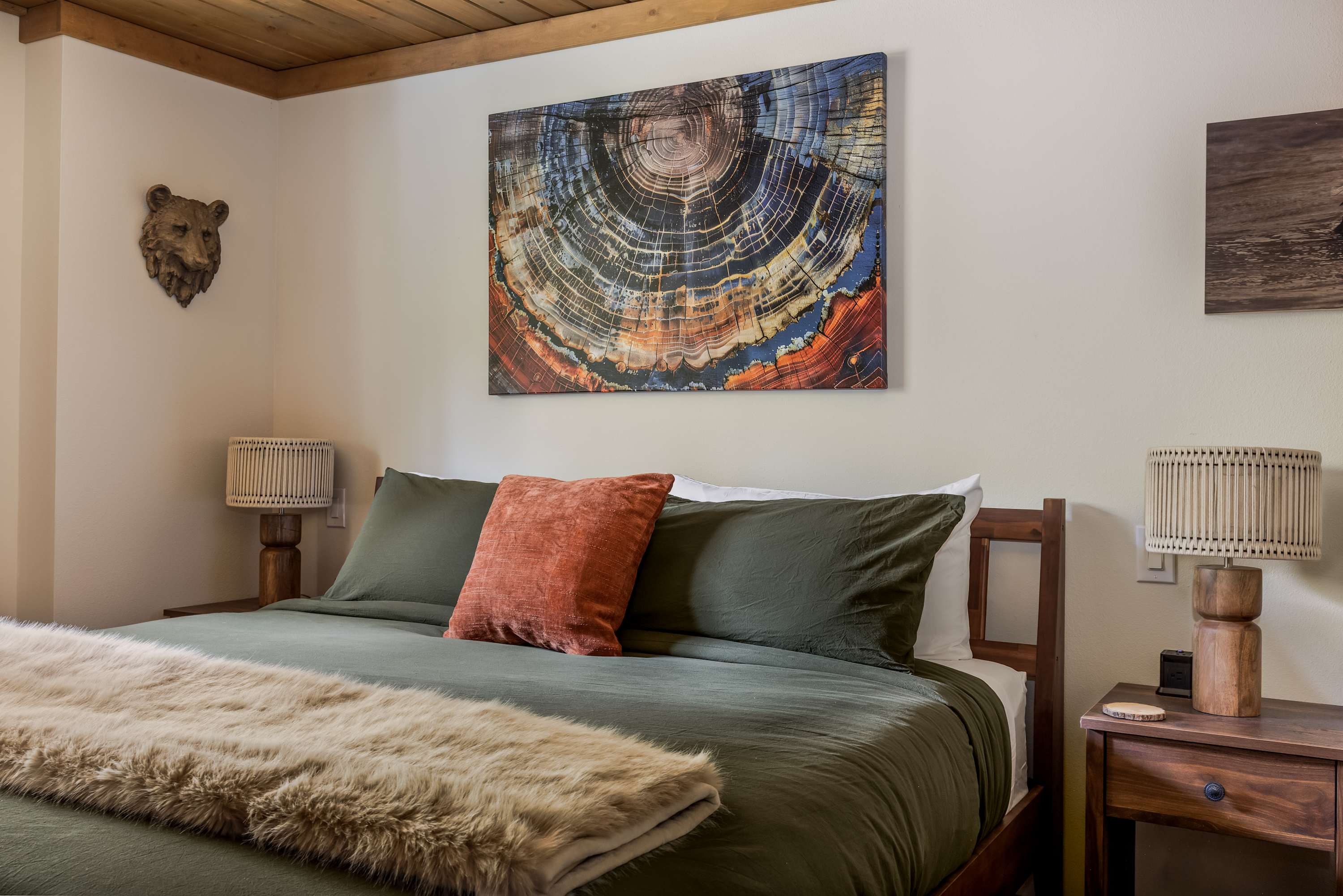 A slightly different perspective of the king bed, showcasing the dark green bedding, orange pillow, and faux fur throw. The wooden plank ceiling and nature-themed artwork, including a prominent tree ring print and a bear head sculpture, contribute to the rustic aesthetic.