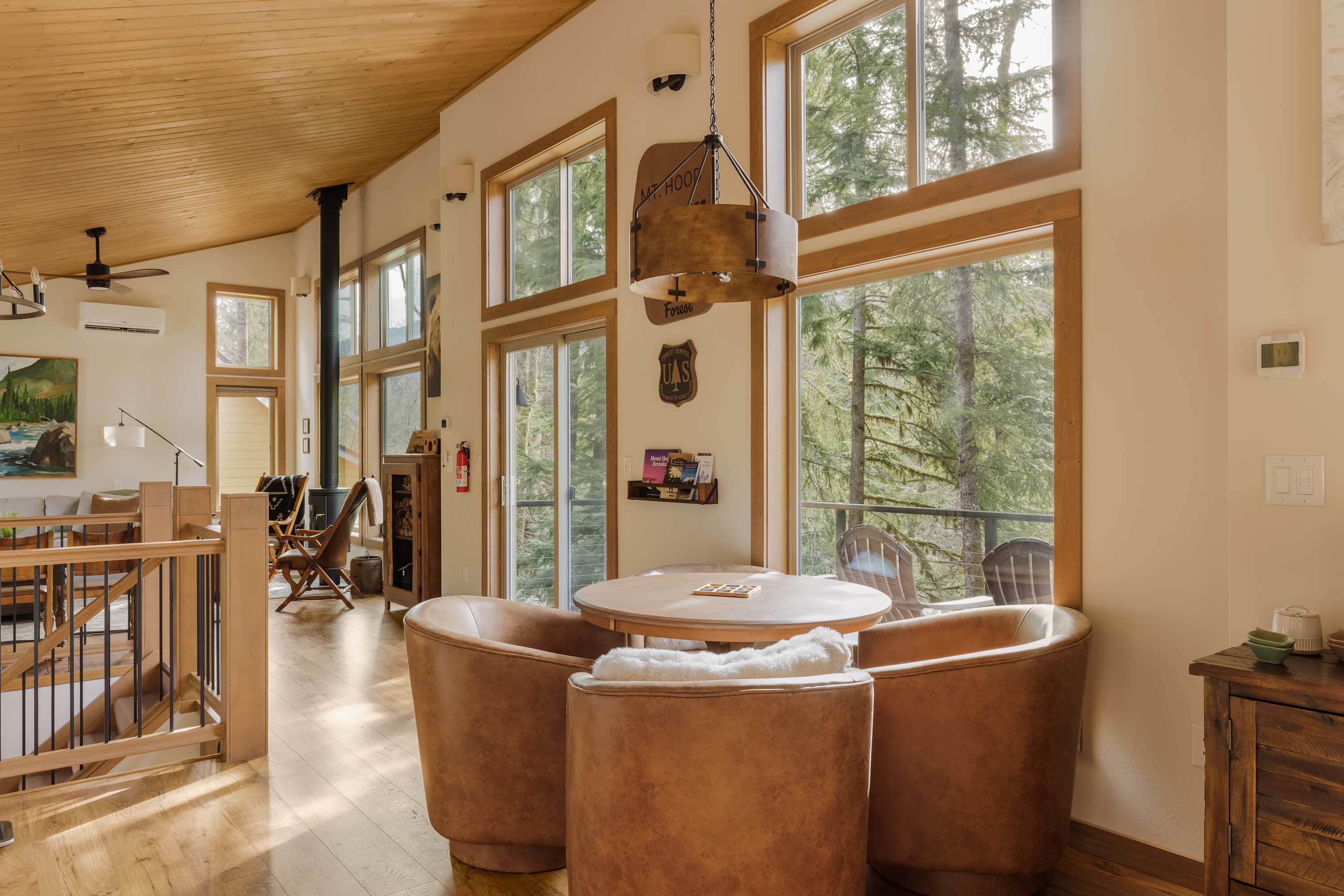 View of the dining area with a round wooden table and four brown leather chairs, overlooking a deck and forest through large windows. The living room with a wood stove is visible in the background.