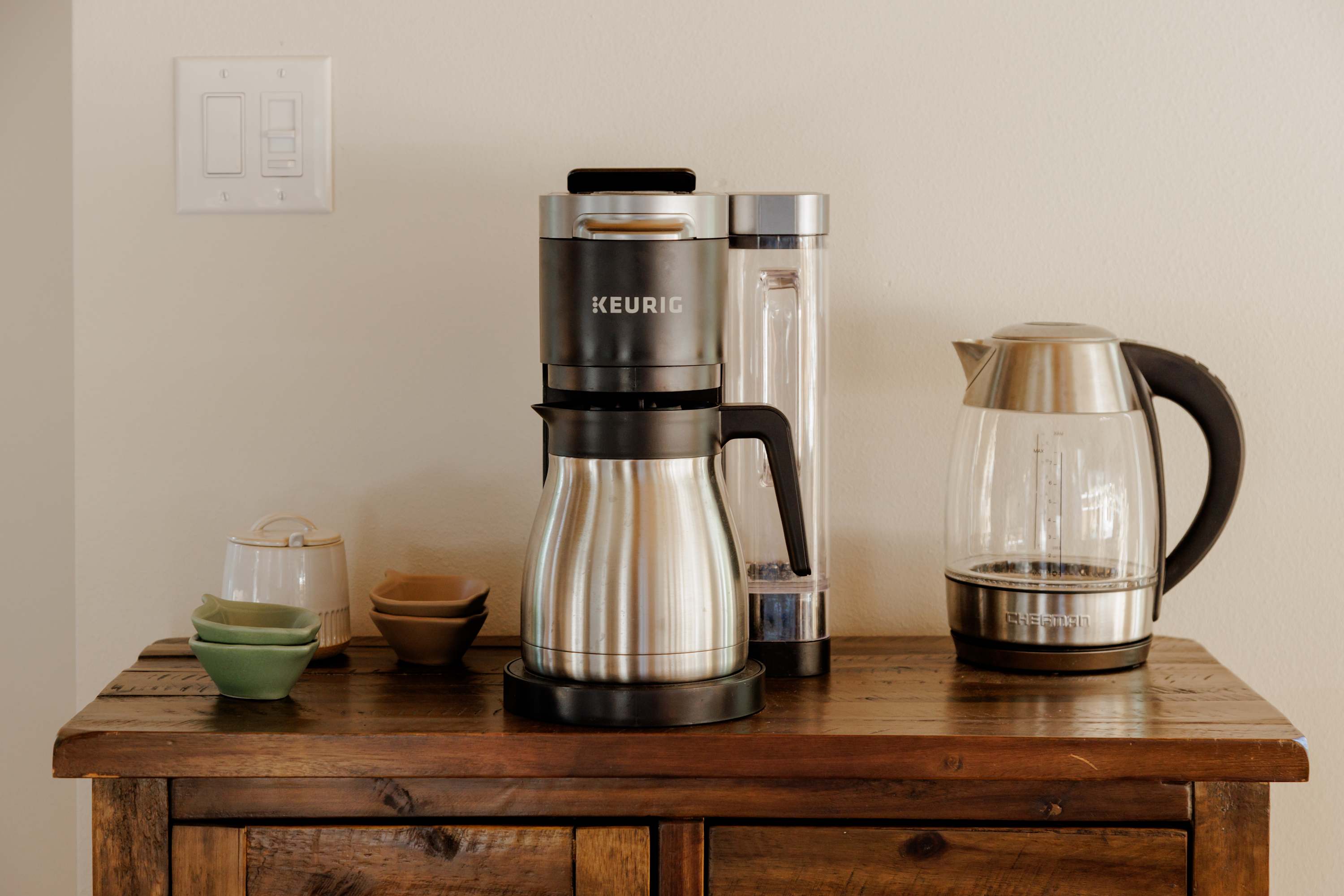 Coffee station area with a Keurig coffee maker and an electric kettle on a rustic wooden cabinet. Small bowls and cups are neatly arranged beside the appliances.