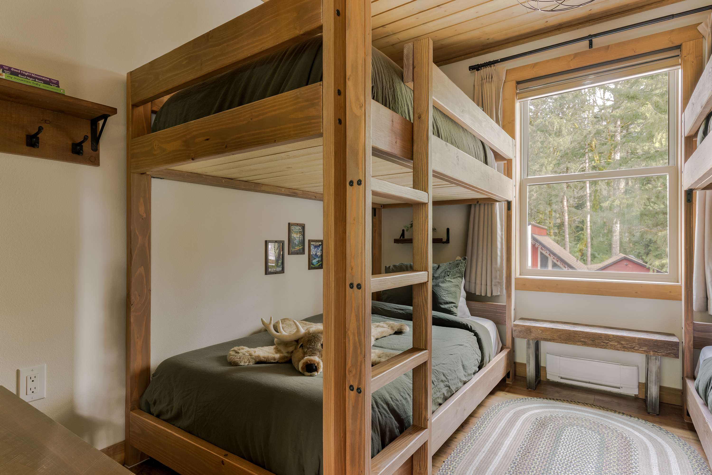 A cozy bunk room featuring a wooden bunk bed with dark green bedding and a plush moose toy. The room has a large window with a view of trees and a wooden plank ceiling.